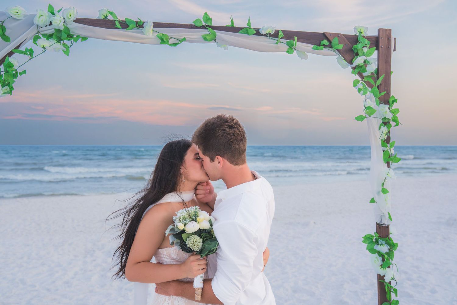 Couple kissing under a wooden arch with green ivy on a sandy beach at sunset, bride holding white roses bouquet.
