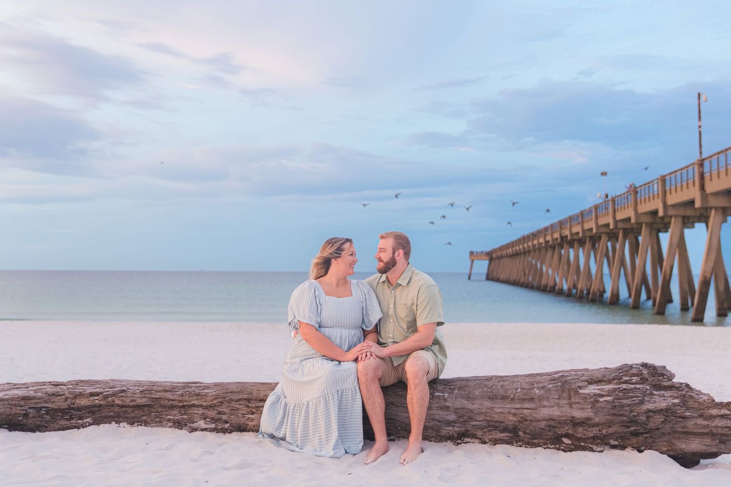 Couple sitting on driftwood log on white sand beach near pier at sunset.