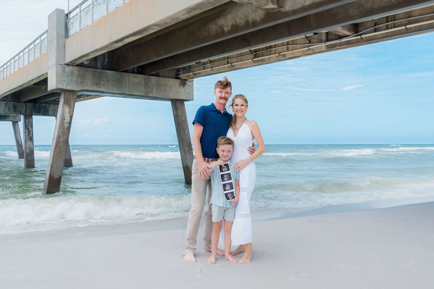 Family of three poses on beach near pier, mom holding ultrasound photos, announcing pregnancy at ocean shore.