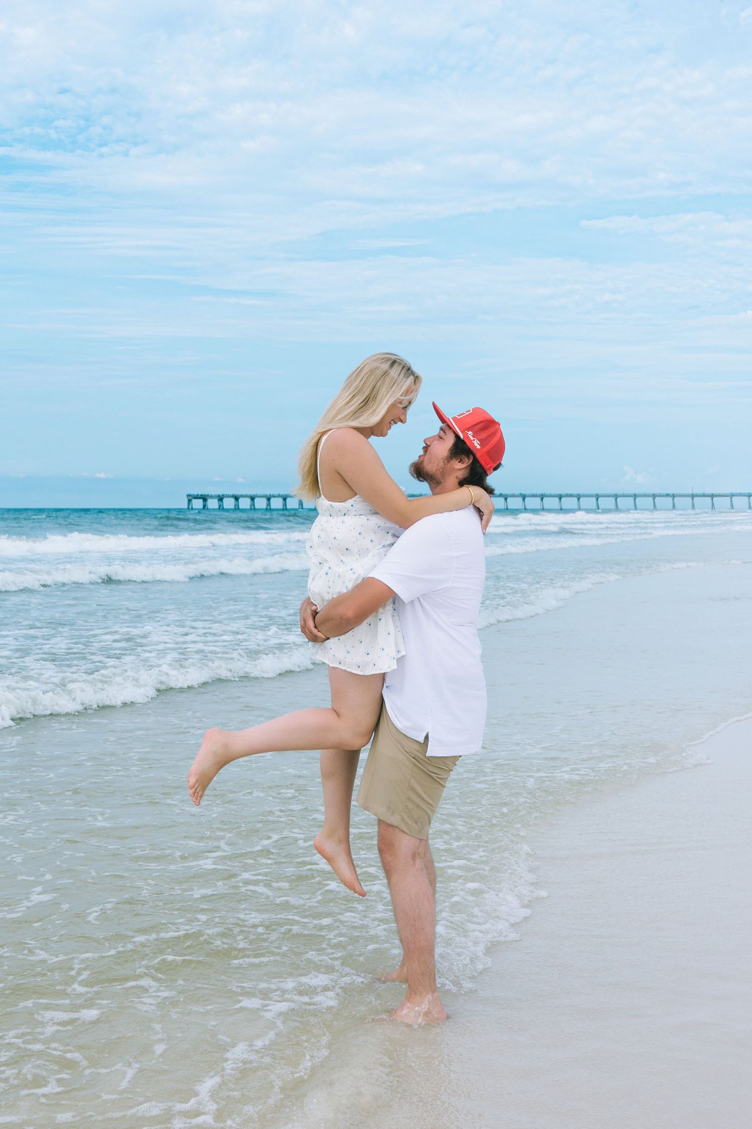 Couple embracing on a sunny beach, man in red cap lifting woman in white dress near ocean waves.