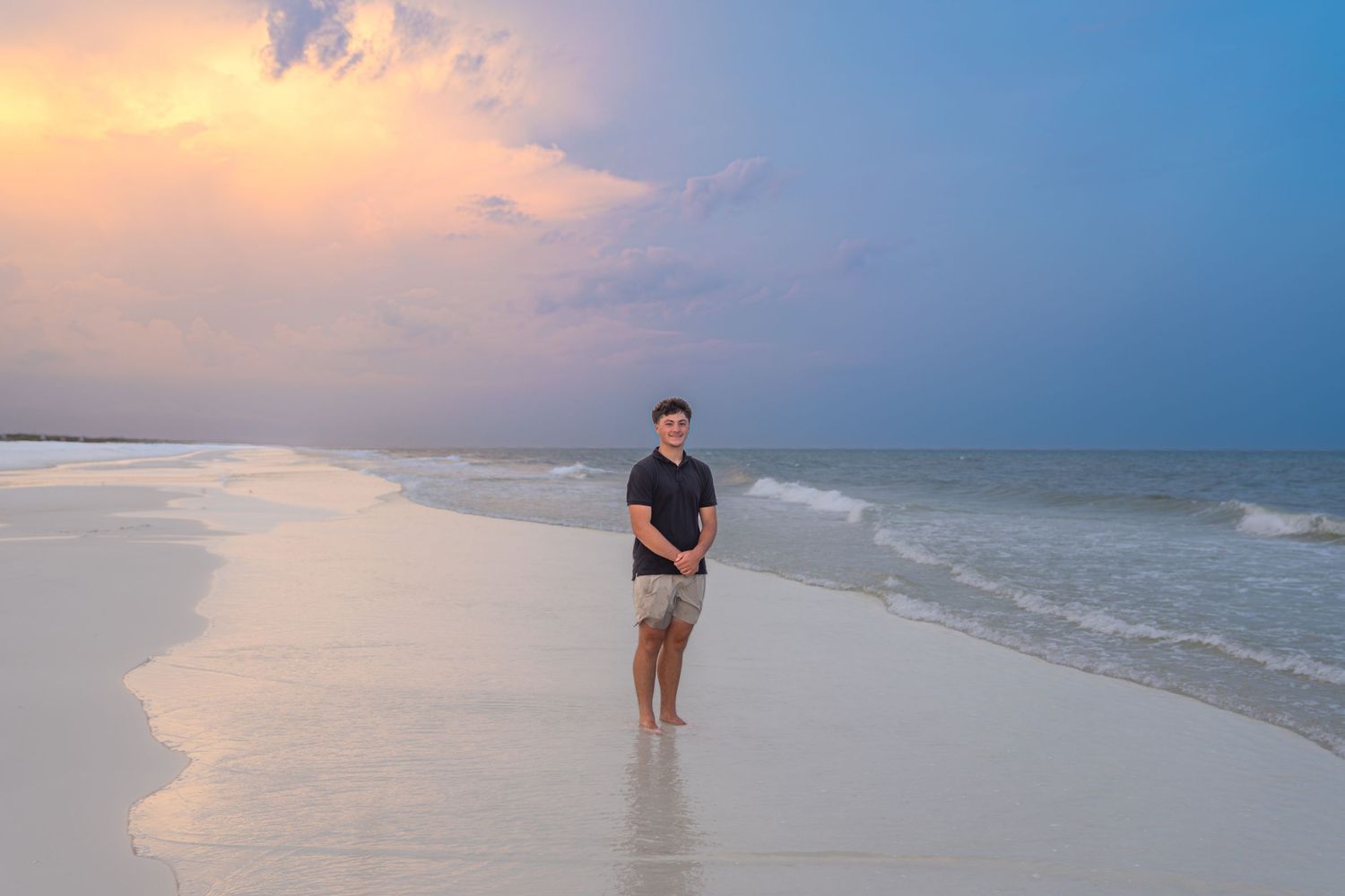 Young man standing barefoot on a sandy beach at sunset with colorful sky and ocean waves in the background.