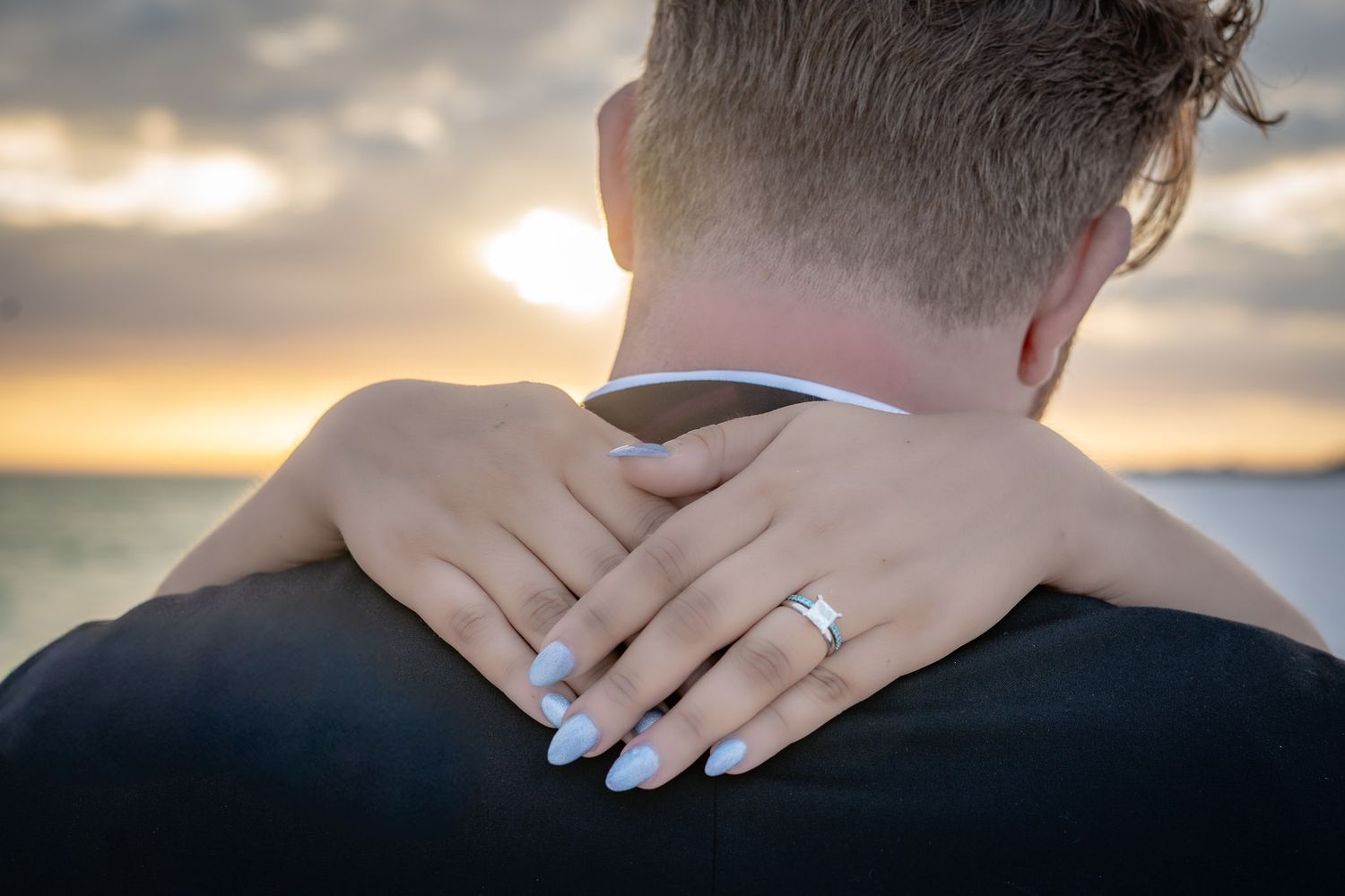 Close-up of bride resting hand on groom's shoulder, showcasing diamond engagement ring at sunset.