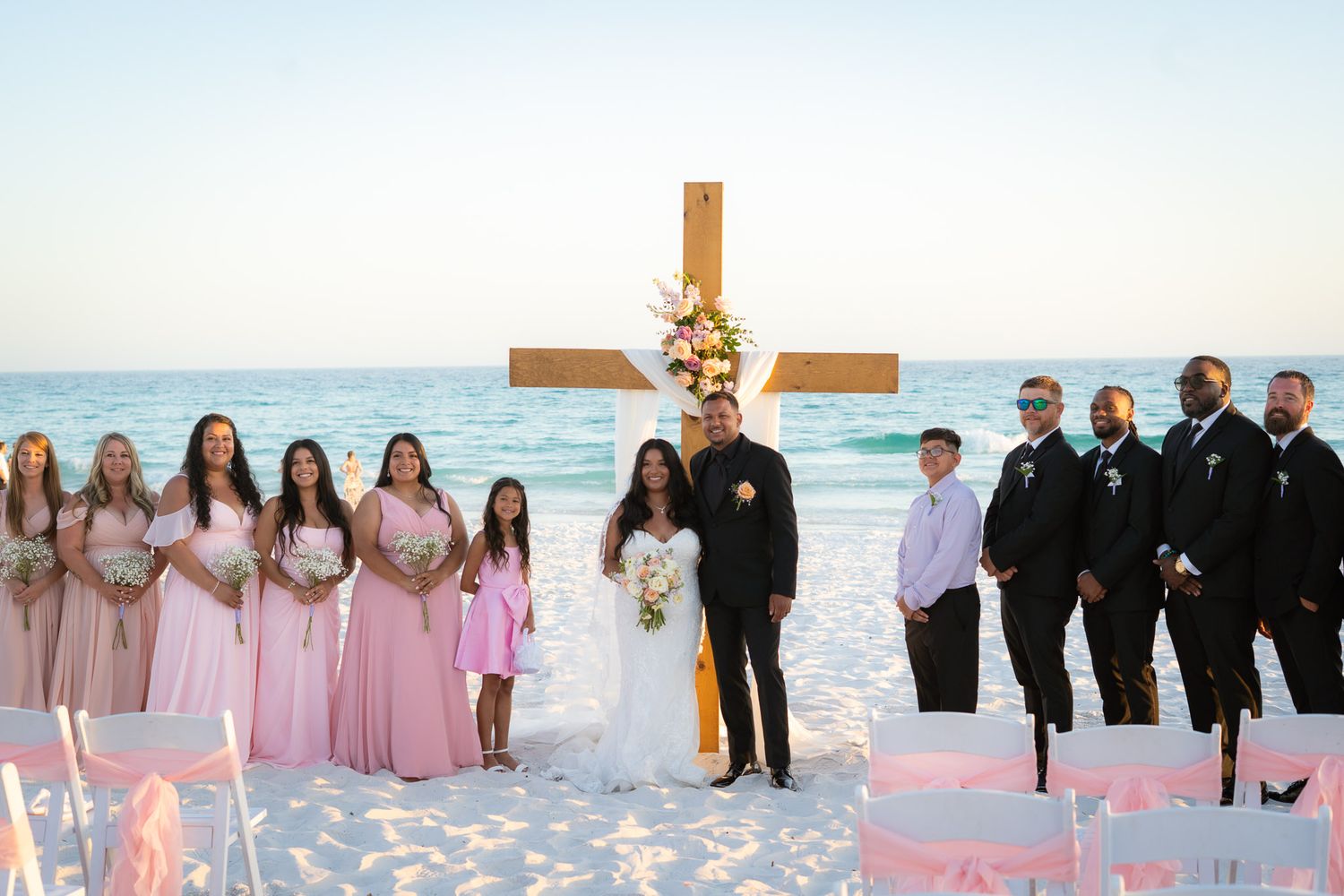 Bride and groom pose with bridesmaids in pink and groomsmen in black suits at a beachfront wedding ceremony.