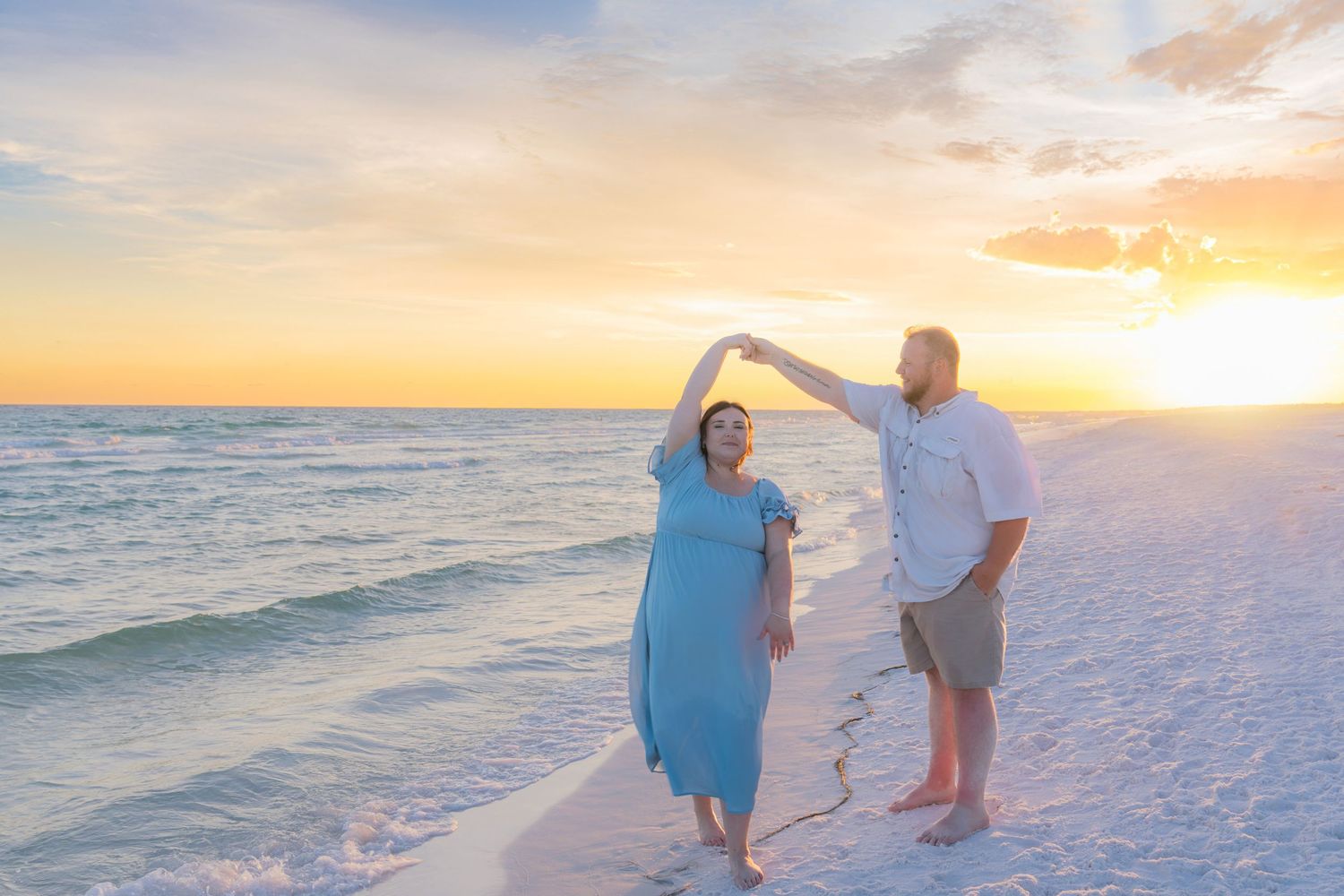 Couple dancing on a white sand beach at sunset with golden skies and calm ocean waves in the background.