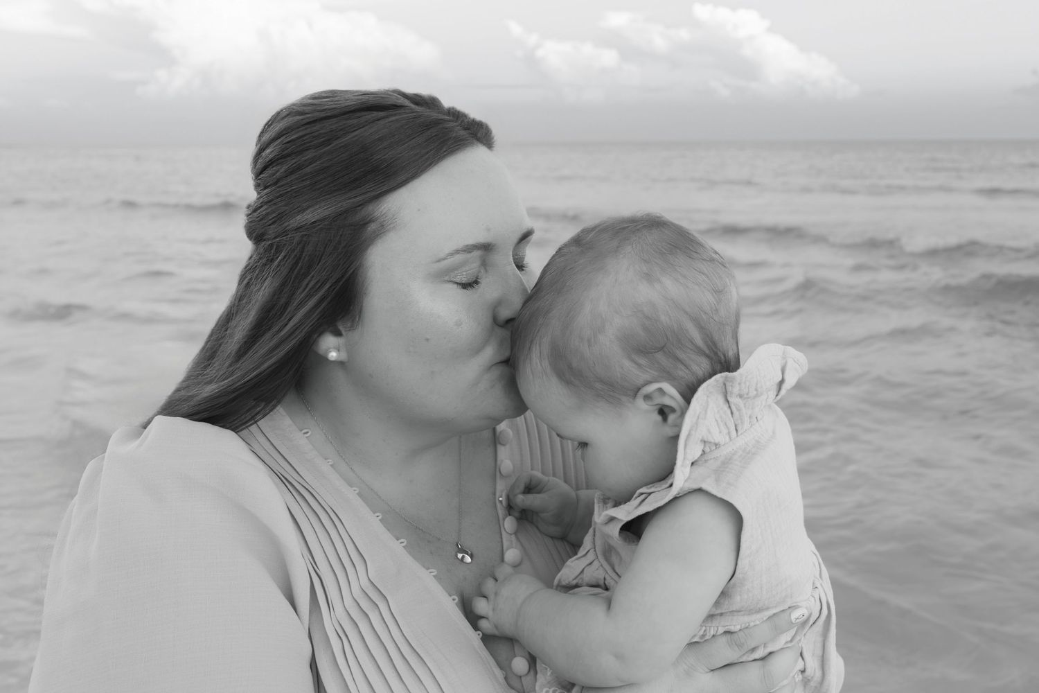 Black and white photo of a mother kissing her baby on the cheek at the beach with ocean waves in the background.