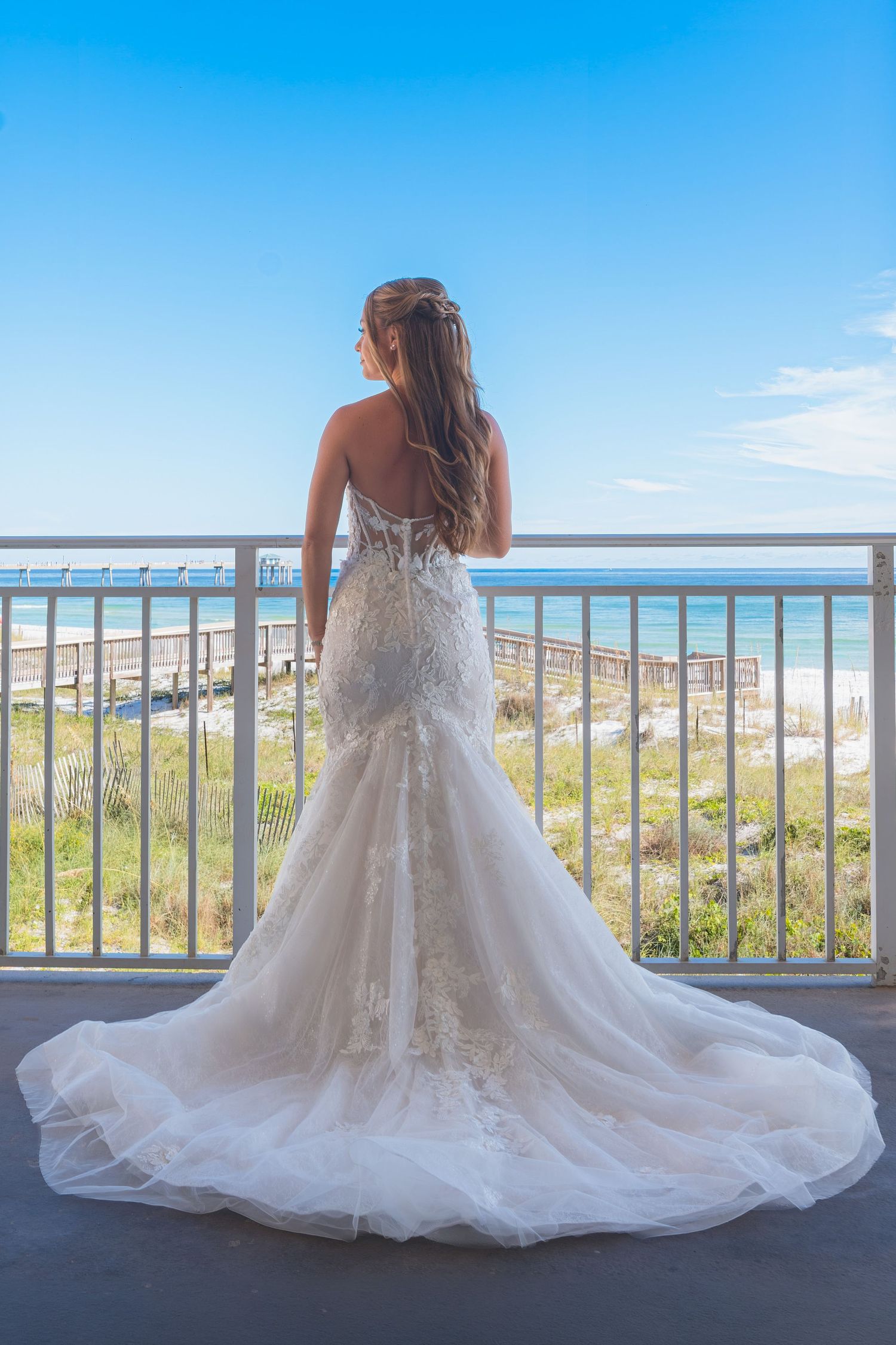 Bride in elegant lace mermaid wedding gown with train stands on beachfront balcony overlooking ocean under blue sky.