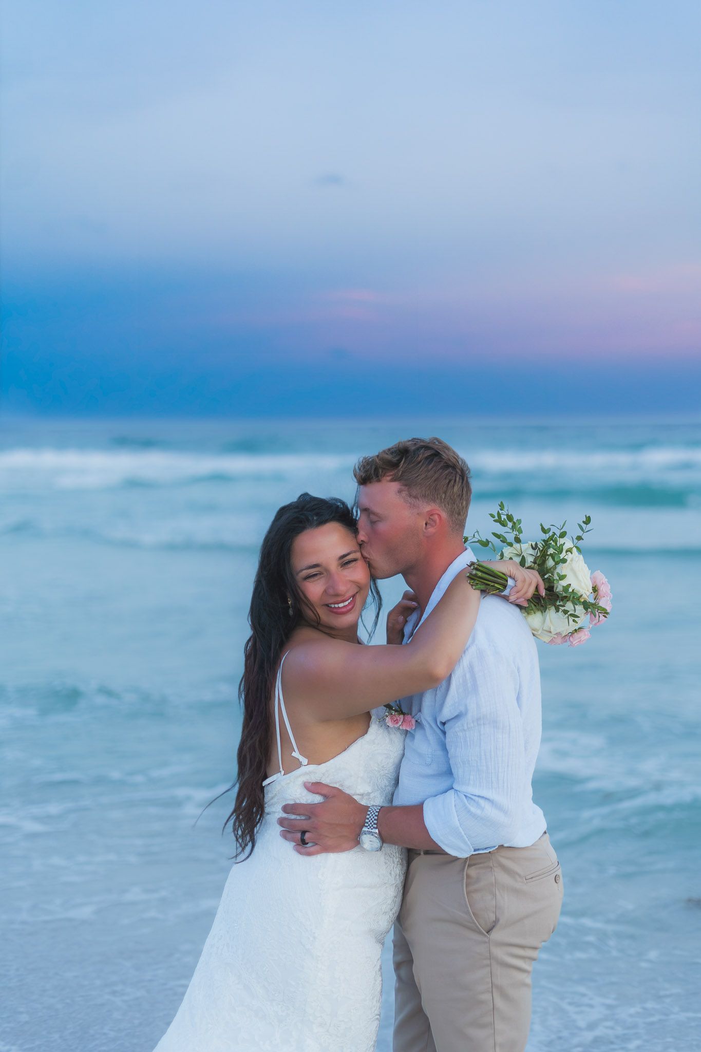 Happy bride and groom embracing on a beach at twilight, groom kissing bride's cheek holding bouquet.