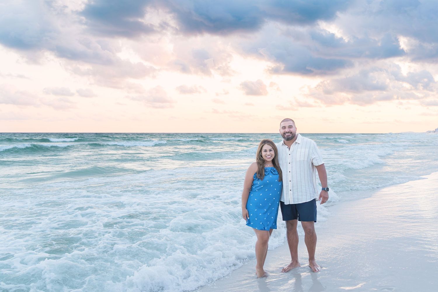 Couple smiling together on a sandy beach at sunset with turquoise waves and colorful clouds behind them.
