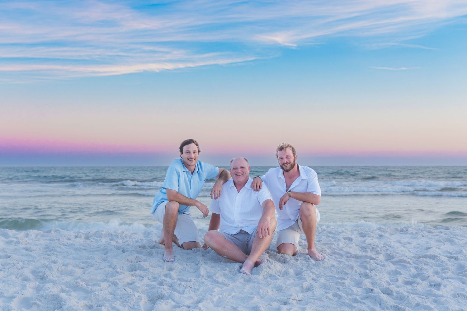 Three men pose together on a white sand beach at sunset with pink and blue sky over calm ocean waters.