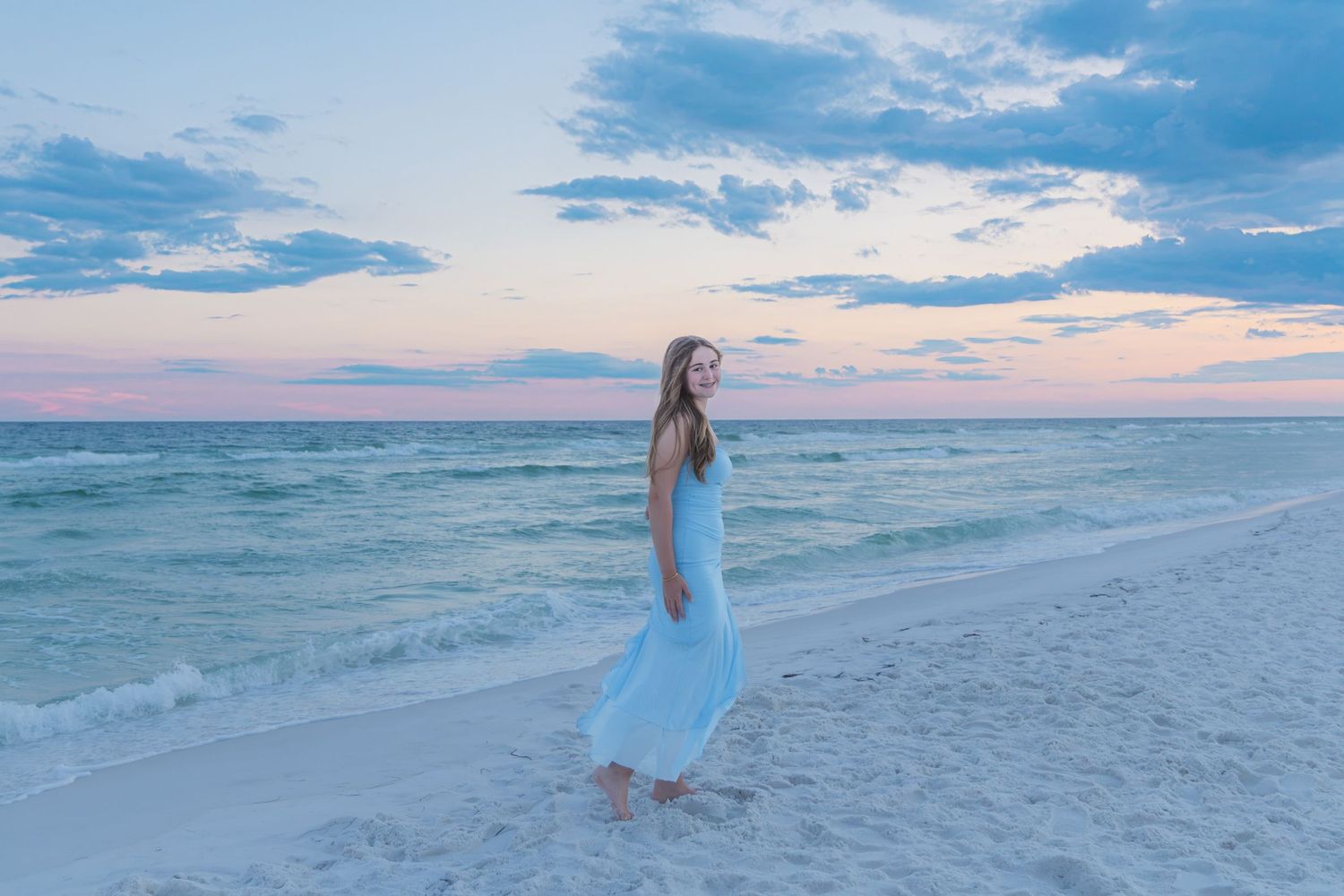 Young woman in flowing light blue dress walking on white sand beach at sunset with pastel sky and ocean waves.