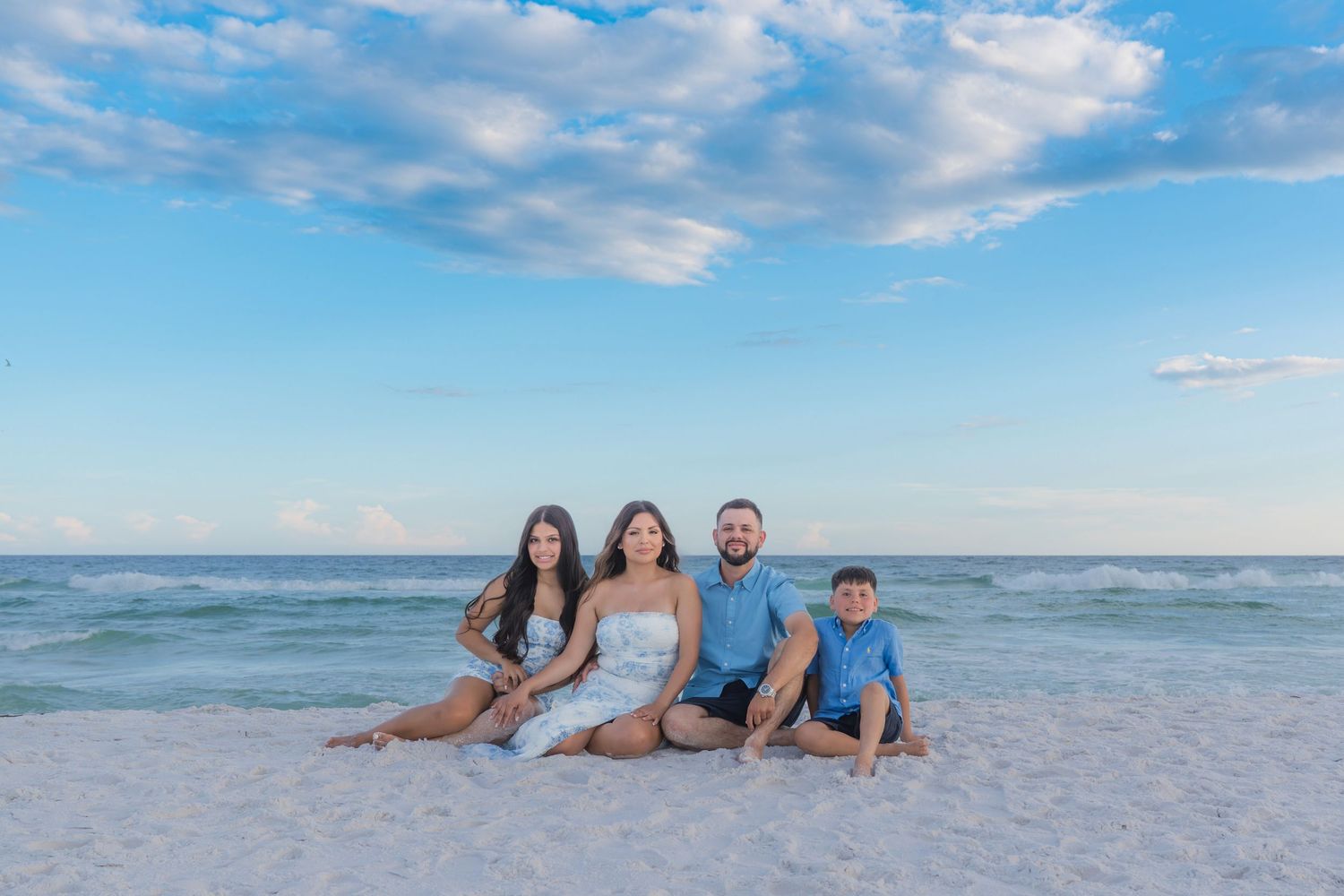 Family of four sitting together on a white sand beach with calm ocean waves and a blue sky backdrop.
