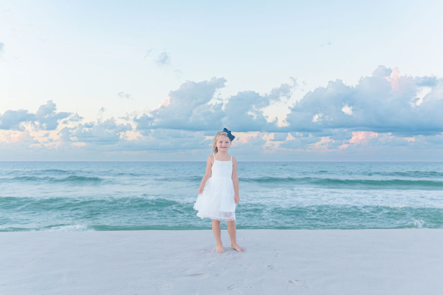 Young girl in white dress stands on white sand beach with turquoise waves and pastel cloudy sky.