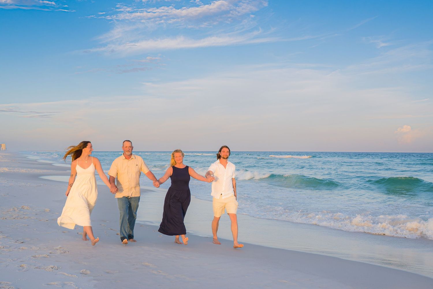 Four people hold hands and walk along a sunny white sand beach with turquoise waves in the background.