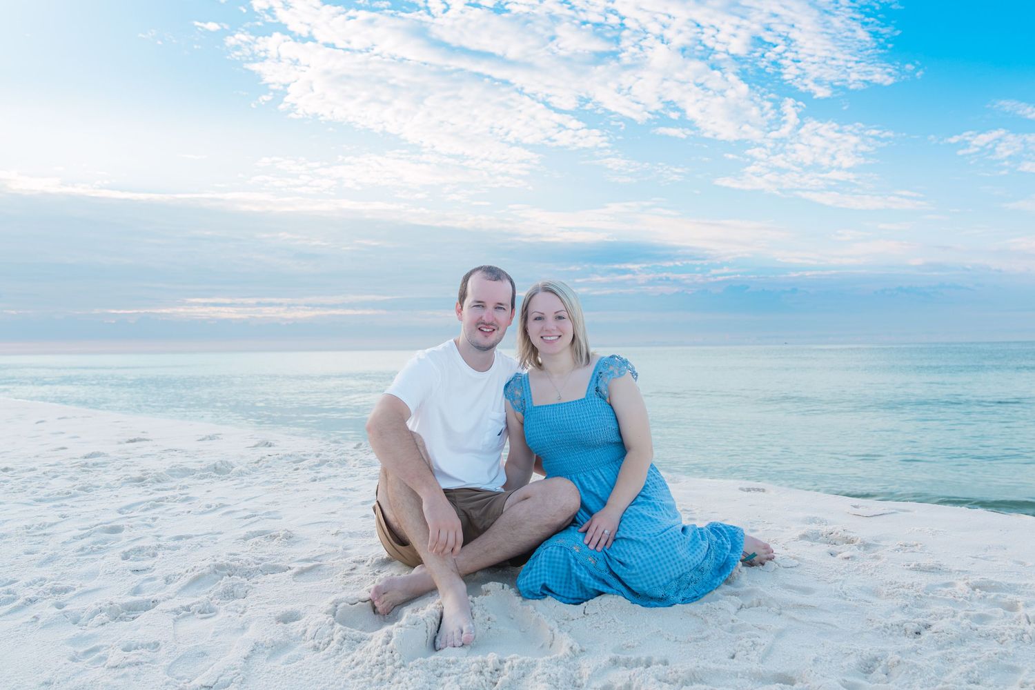 Couple sitting together on white sand beach at sunset, woman in blue dress, man in white shirt, ocean backdrop.