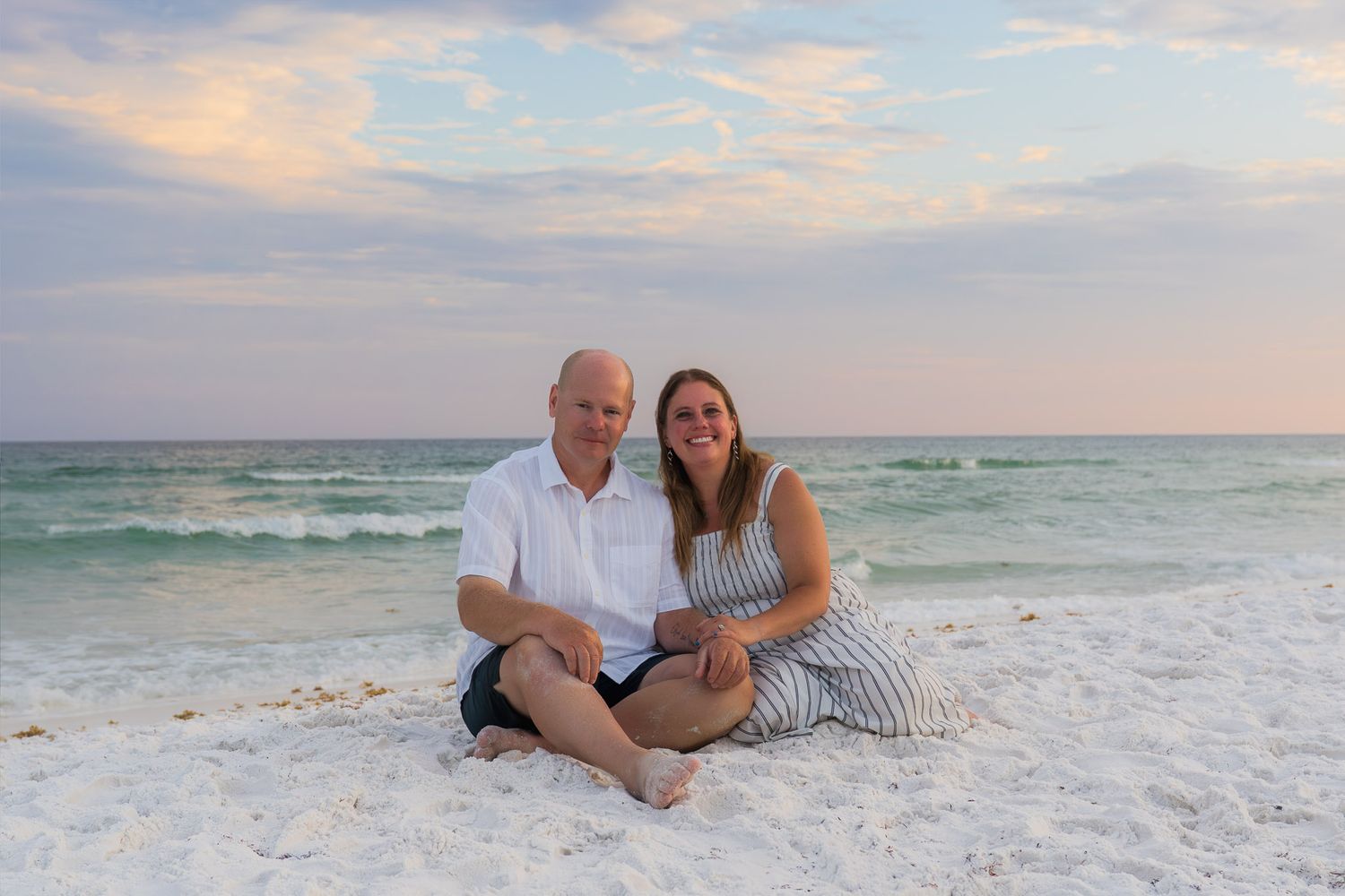 Couple sitting together on white sand beach at sunset with turquoise ocean waves in background.