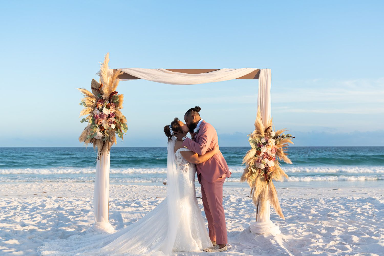 Couple kissing at beach wedding altar with floral arrangements on white sand with ocean backdrop.