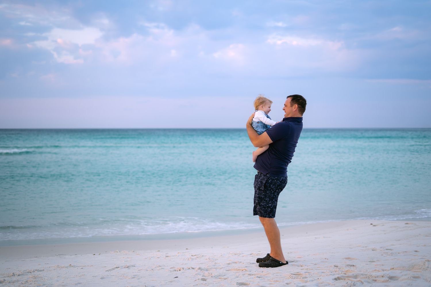 A father holds his baby up on a white sand beach with turquoise ocean water in the background.