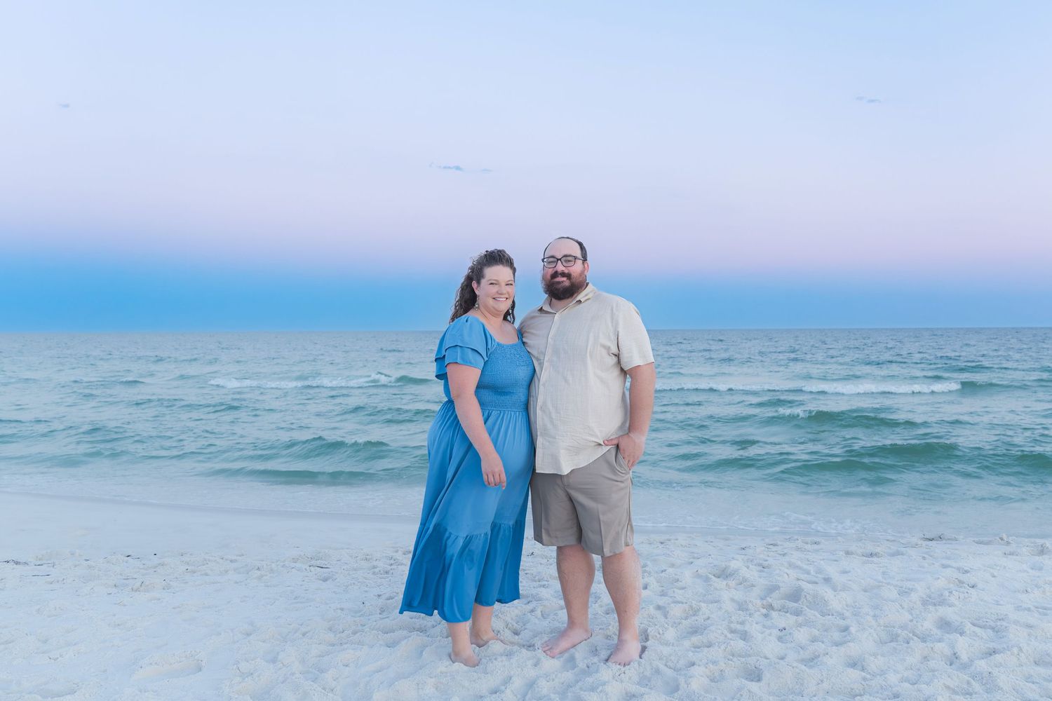 A smiling couple poses together on a white sand beach at sunset, woman in blue dress, man in khaki shirt.