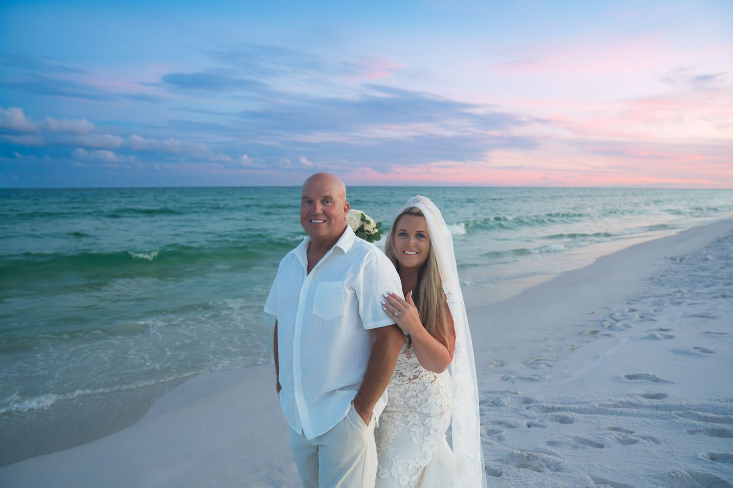 Bride and groom posing on a sandy beach at sunset with turquoise ocean waves and colorful pink sky.
