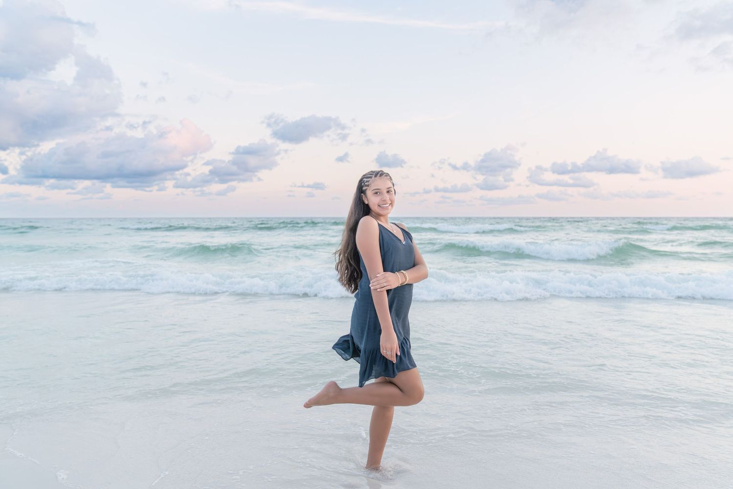 Young woman in blue dress posing gracefully on a sandy beach with turquoise waves and pastel sky.