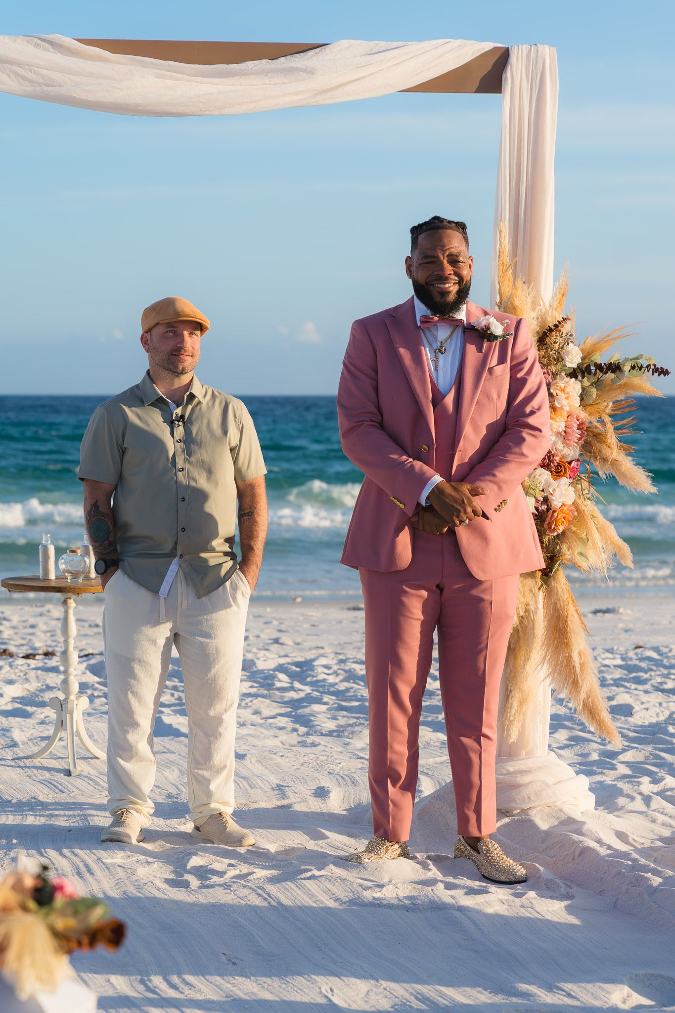 A groom in a pink suit stands beside an officiant at a decorated beach wedding arch with floral arrangements and ocean backdrop.