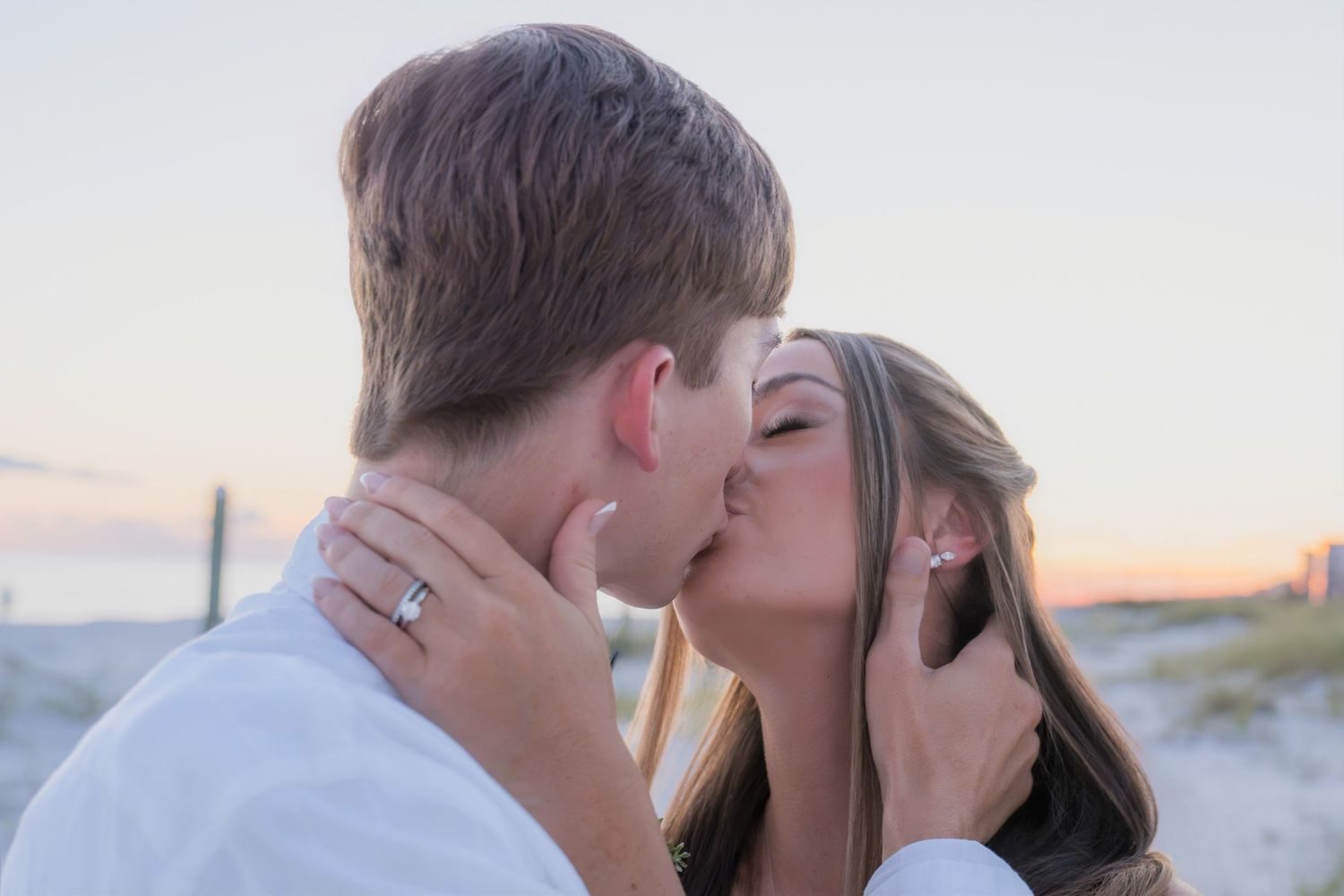 Couple sharing a romantic kiss on the beach at sunset, woman wearing an engagement ring.