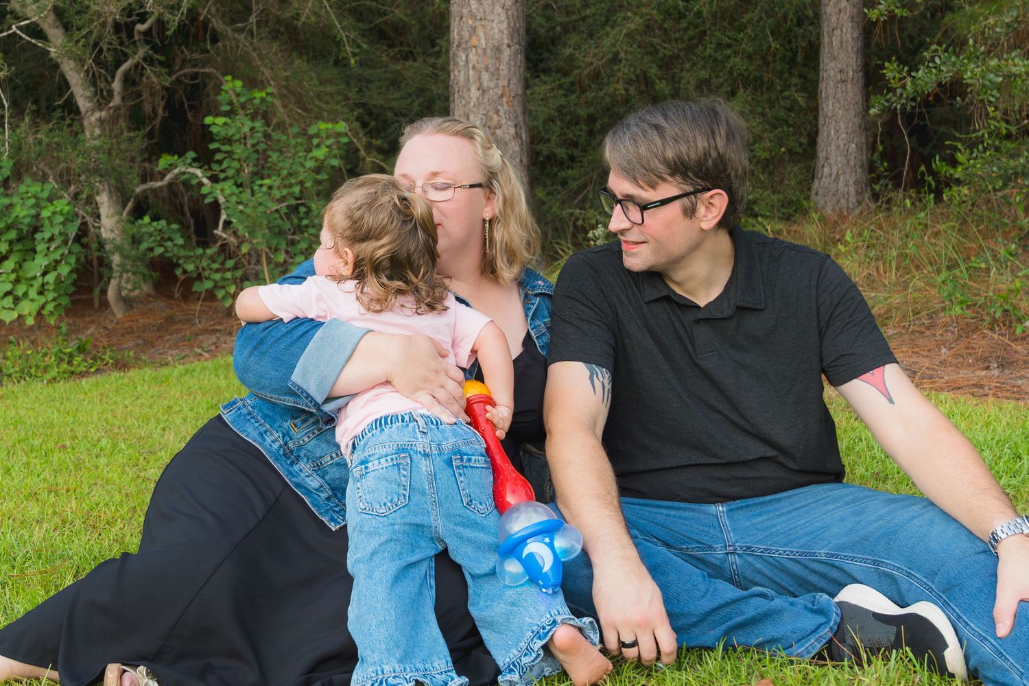 A happy family sits together on grass in a park, with a toddler hugging her mother while father looks on.