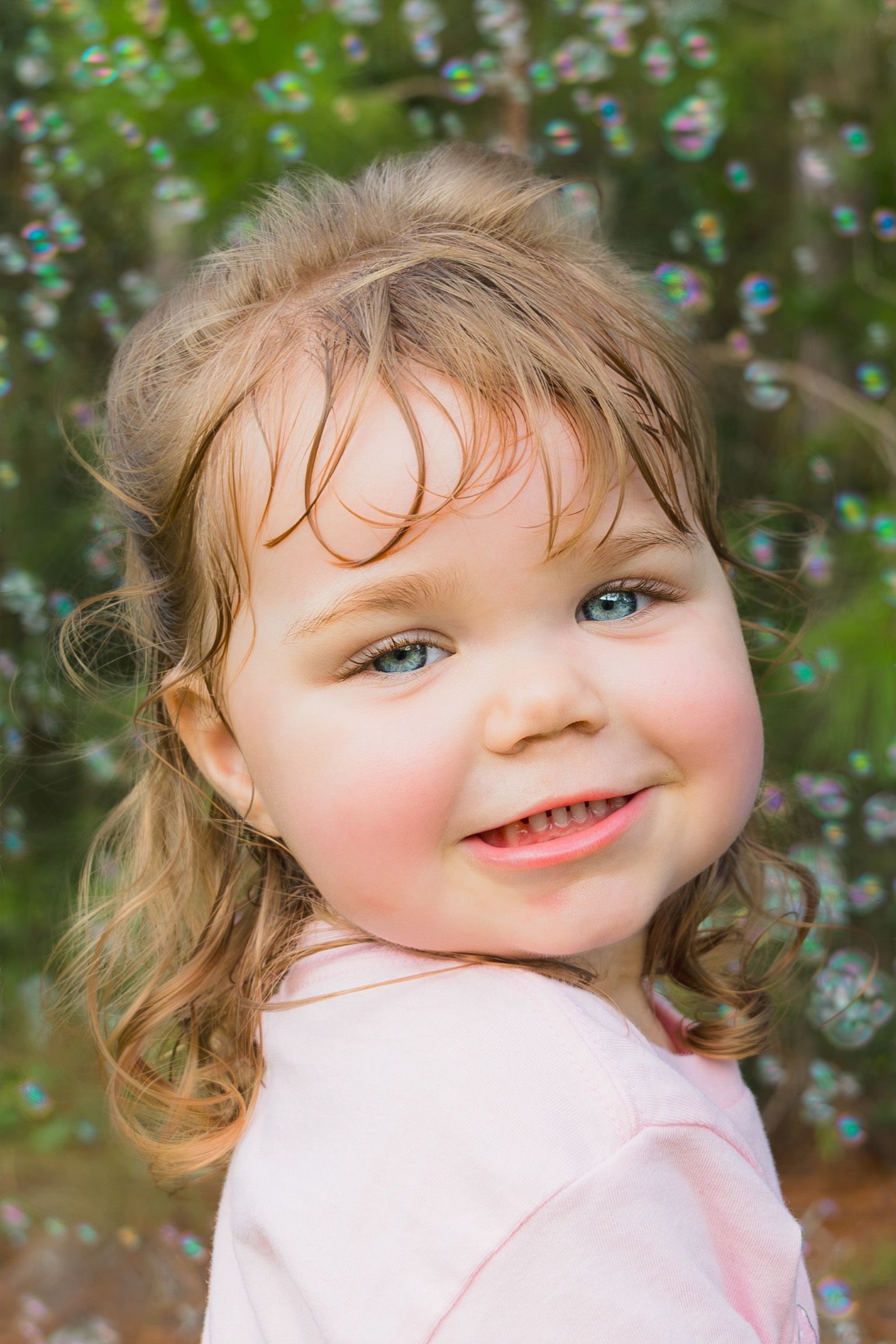Smiling toddler girl with blue eyes and curly hair surrounded by soap bubbles outdoors in a pink shirt.