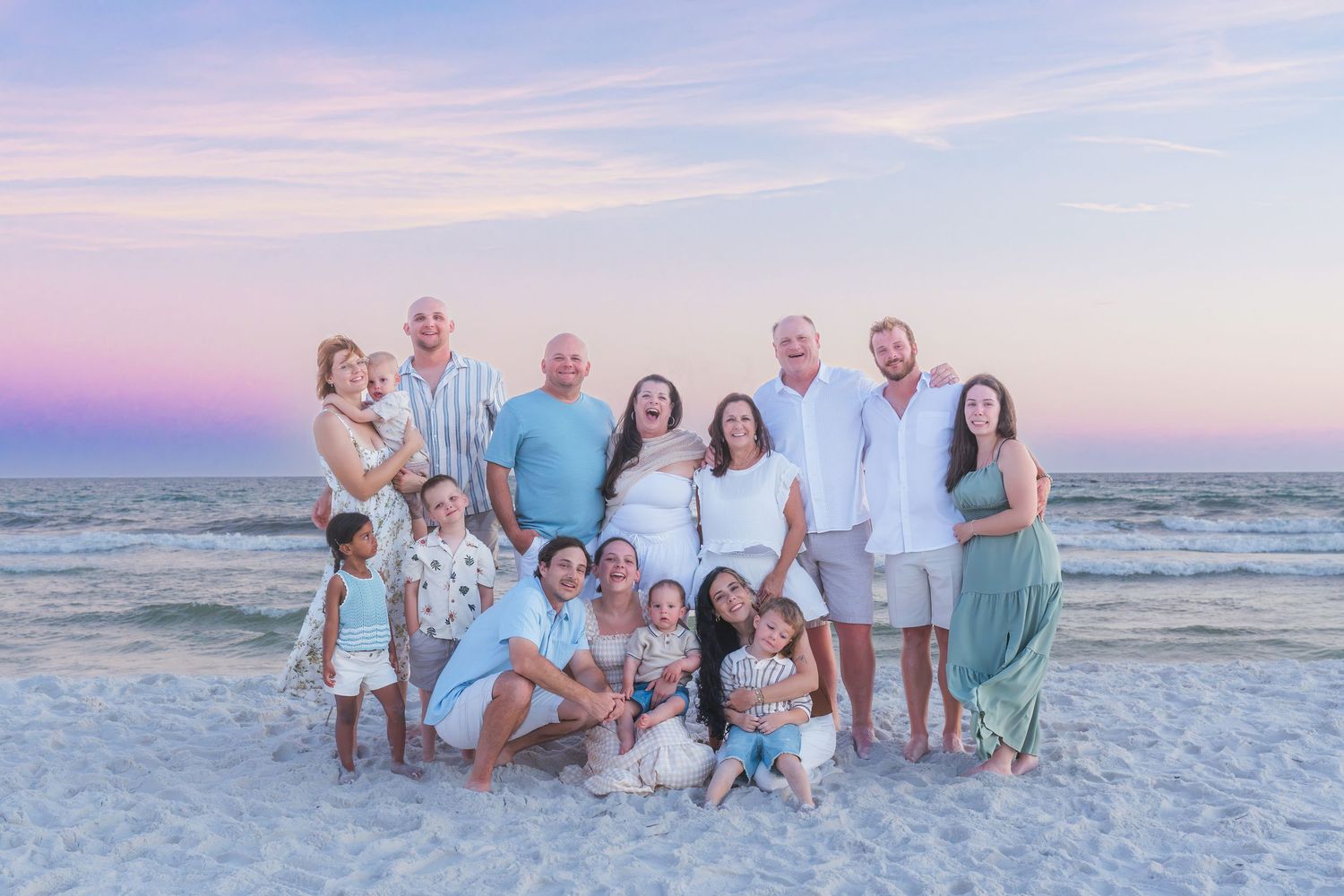 Large family group photo on a sandy beach at sunset with pink and purple sky over calm ocean waters.