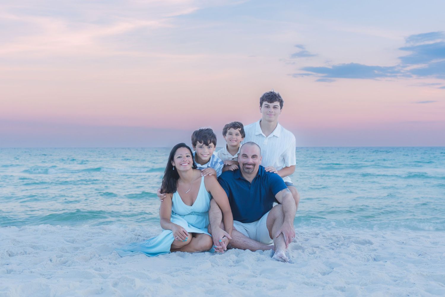 Family of five posing together on a white sand beach at sunset with pastel pink and blue skies over calm turquoise water.