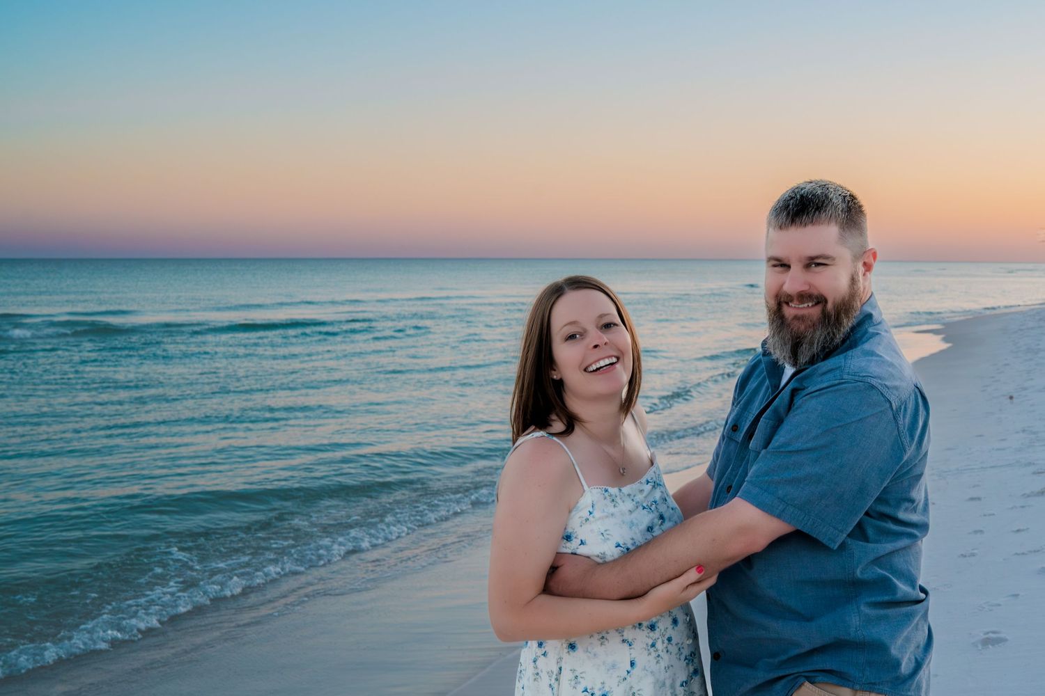 A smiling couple poses together on a sandy beach at sunset with colorful pink and blue skies.