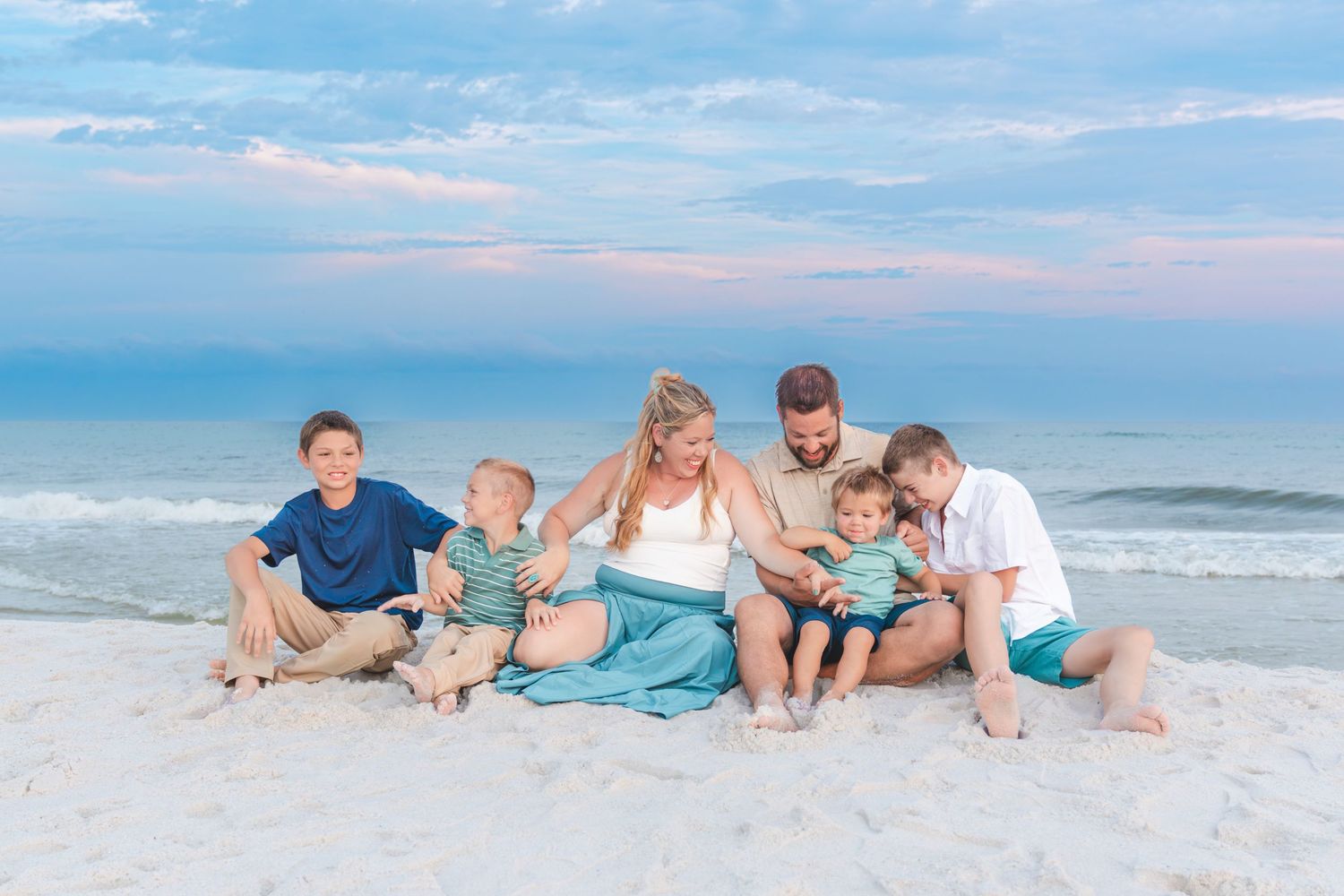 Family of six sitting together on a white sand beach at sunset with calm ocean waves in the background.