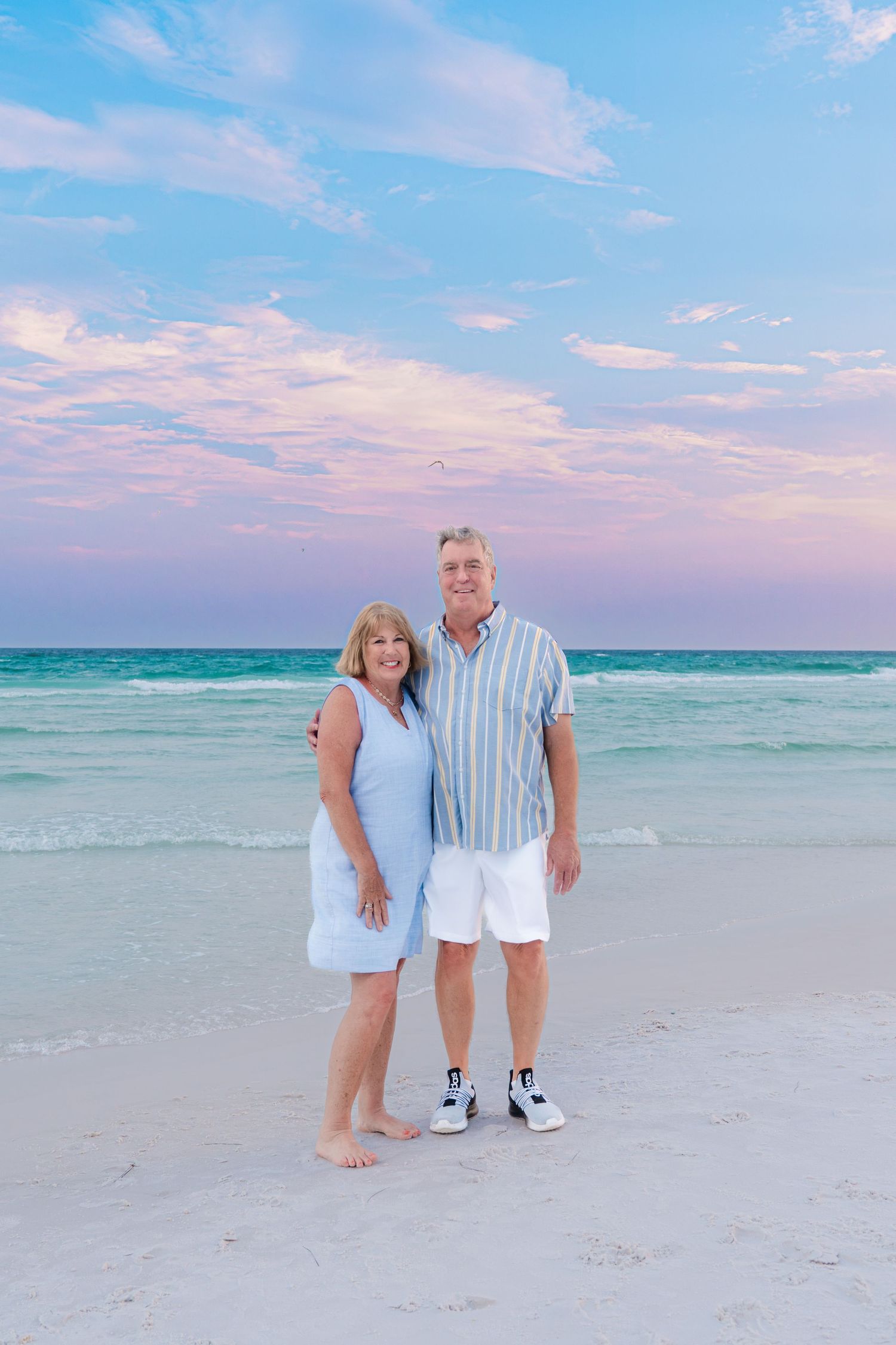 Smiling couple posing on a white sand beach at sunset with turquoise water and pink clouds behind them.