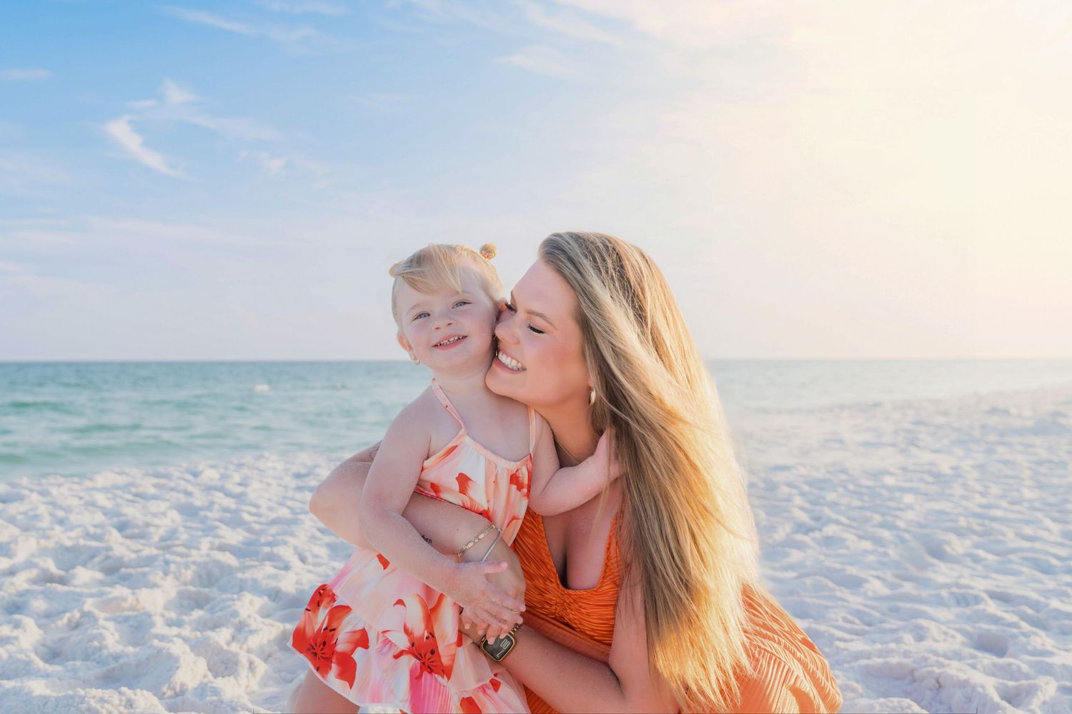 A mother in an orange dress holds her young daughter on a sunny white sand beach with turquoise ocean water.