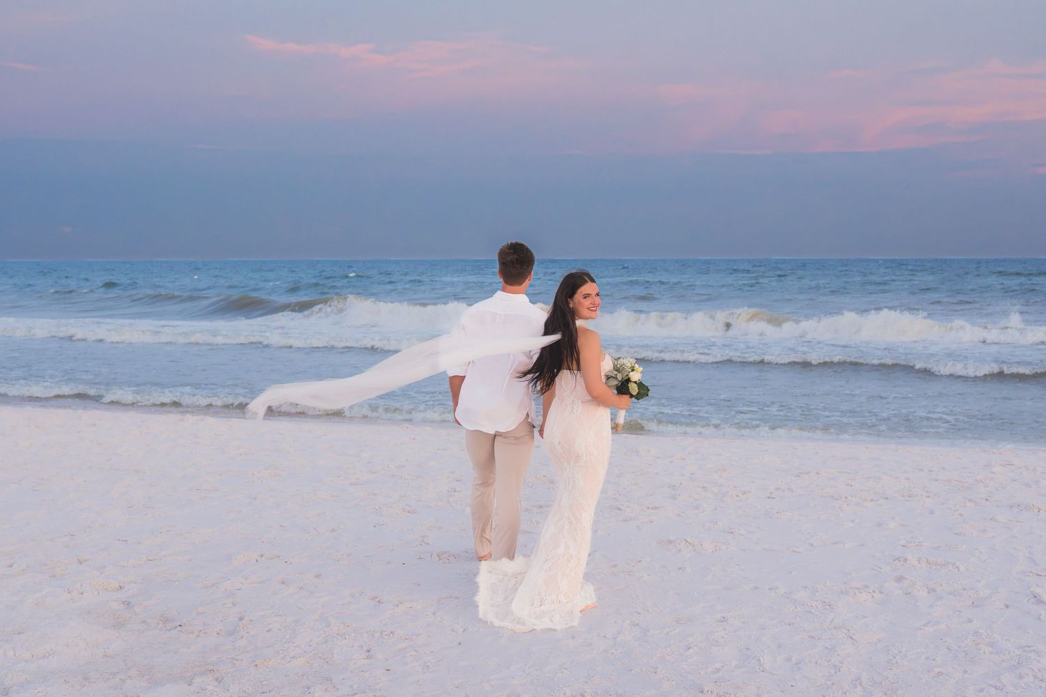 Bride and groom stand back-to-back on a white sand beach at sunset, waves crashing behind them.