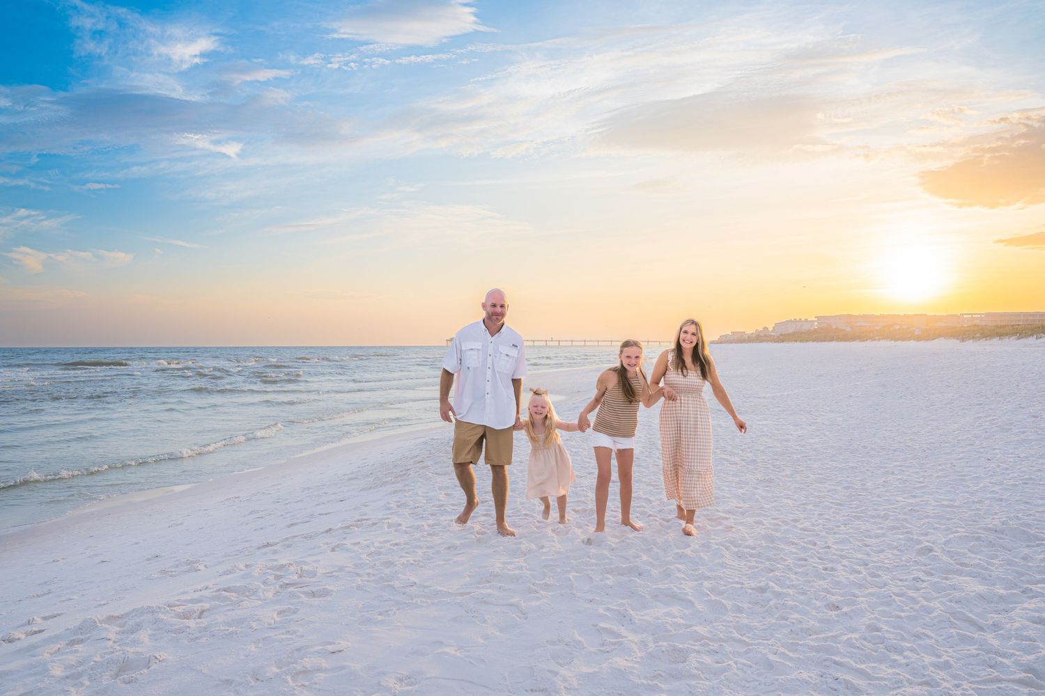 Family of four holding hands walking on white sand beach at golden sunset with calm ocean waves.