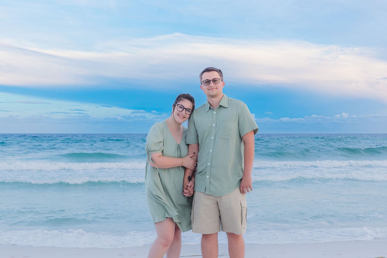 Couple in matching green outfits posing together on a sandy beach with turquoise ocean waves and pastel sky.