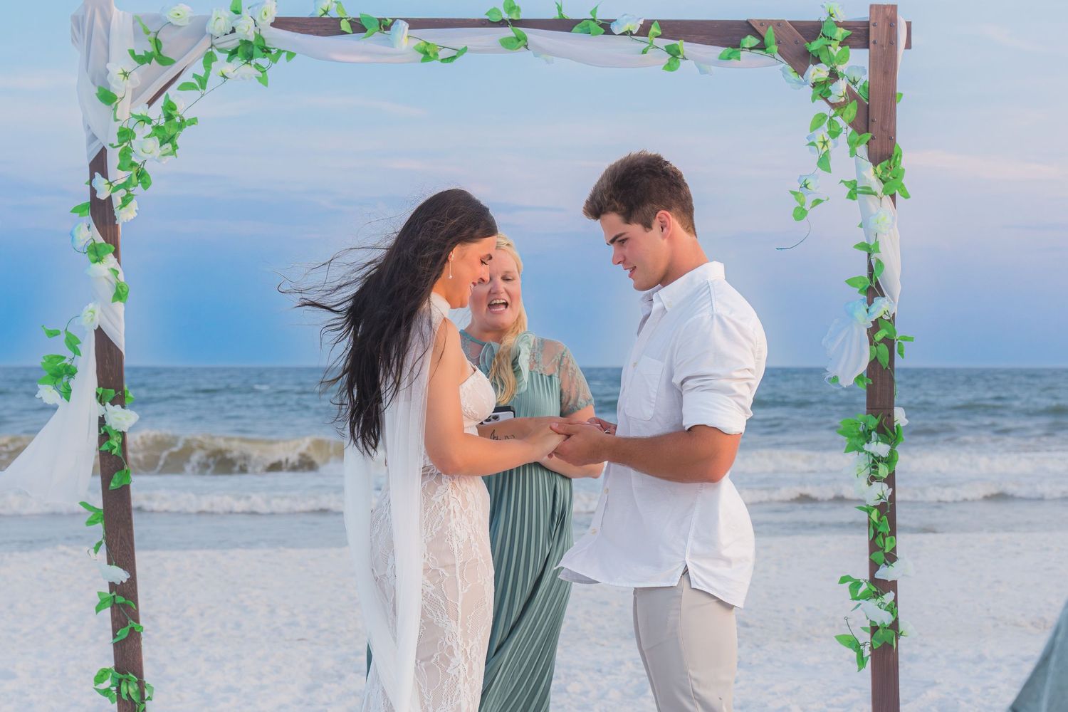 Couple exchanges vows at a romantic beach wedding ceremony under a floral arch with ocean backdrop.