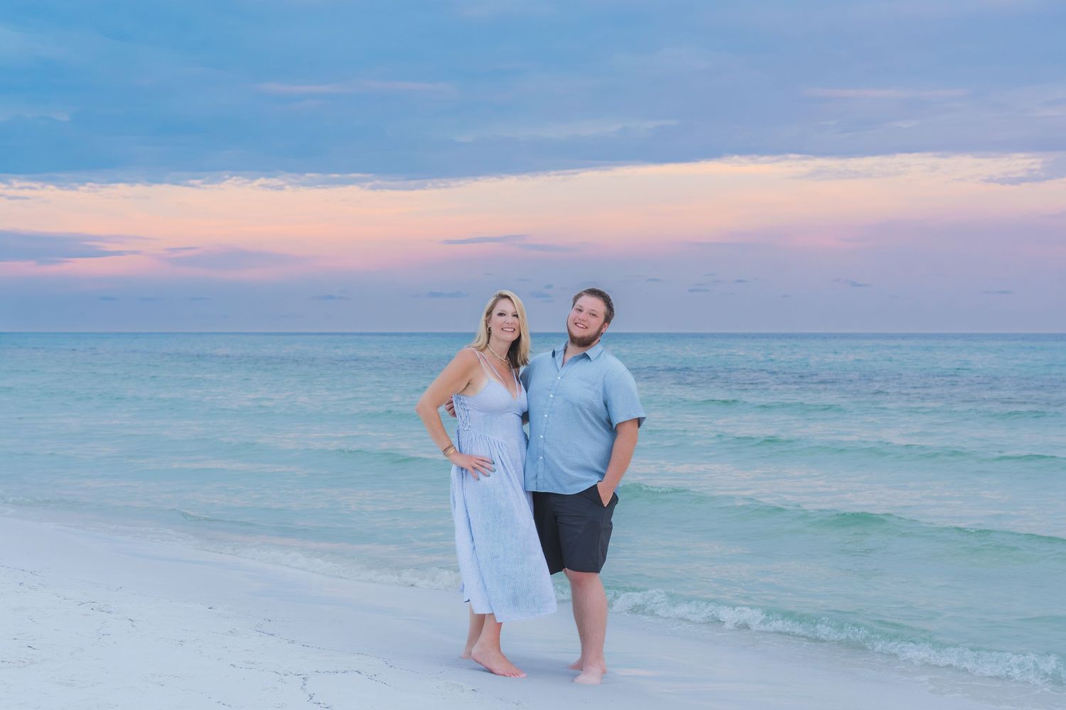 Couple posing together on a white sand beach at sunset with pastel pink and blue sky over turquoise water.