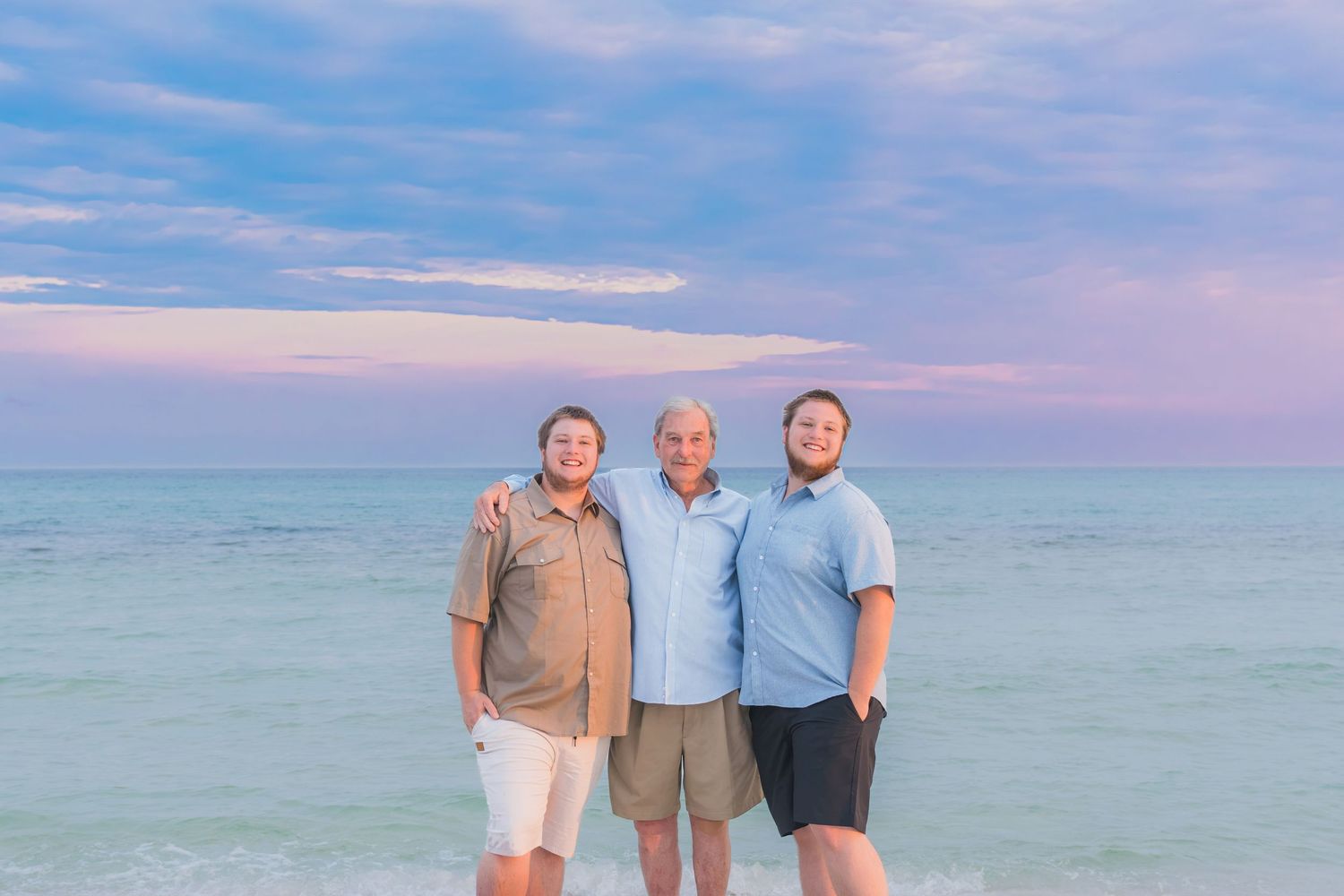 Three men smiling together on a beach at sunset with calm ocean waters and colorful sky behind them.
