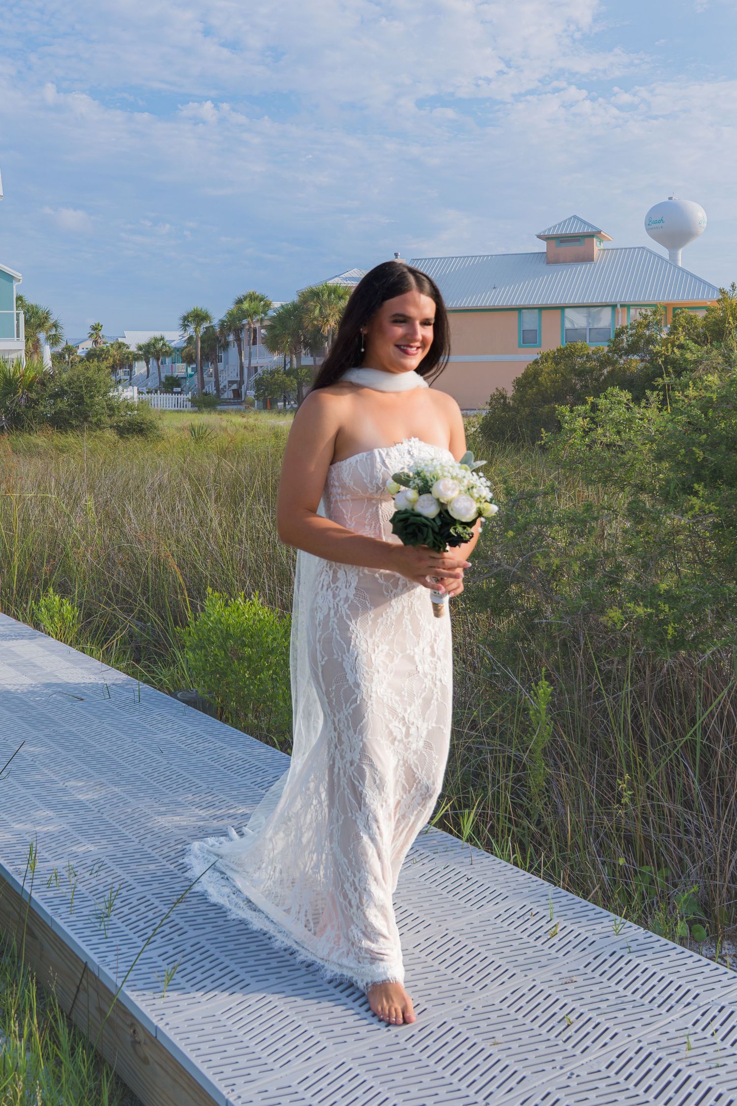 Smiling bride in strapless white lace gown holds bouquet on boardwalk near beach dunes and resort buildings.