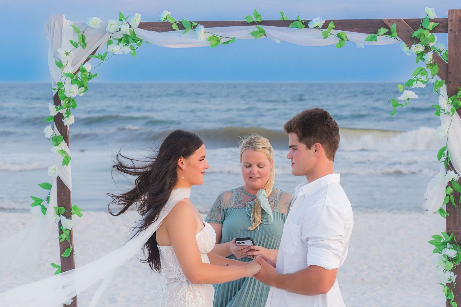 Couple exchanging vows at a beach wedding ceremony under a floral arch with an officiant by the ocean.