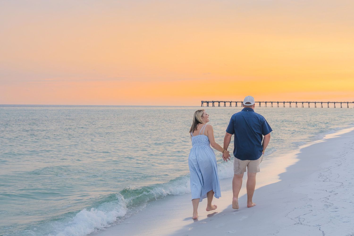 Couple holding hands walking on white sand beach at sunset with pier in background.