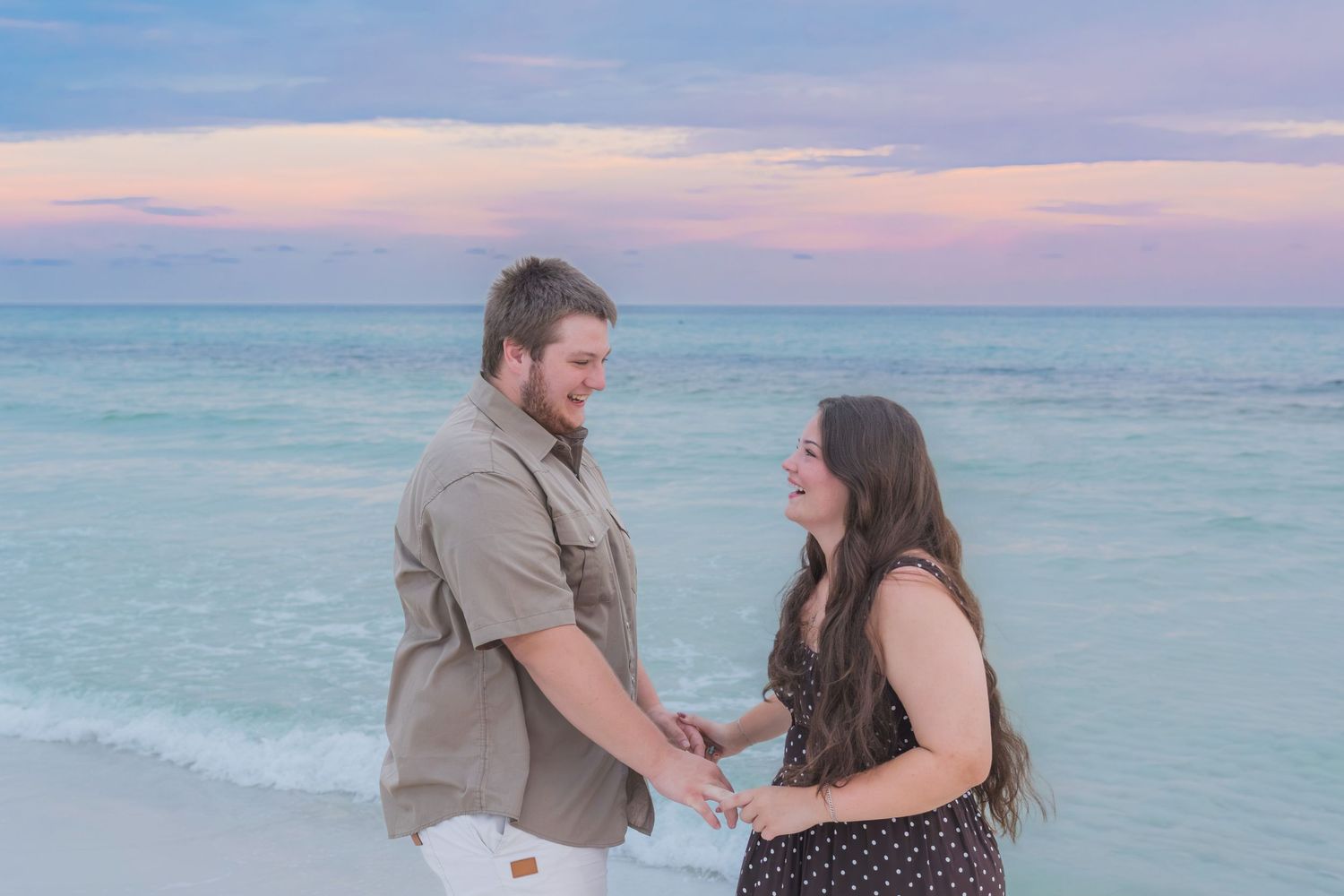 Couple holding hands on a beach at sunset with turquoise water and pink sky in the background.
