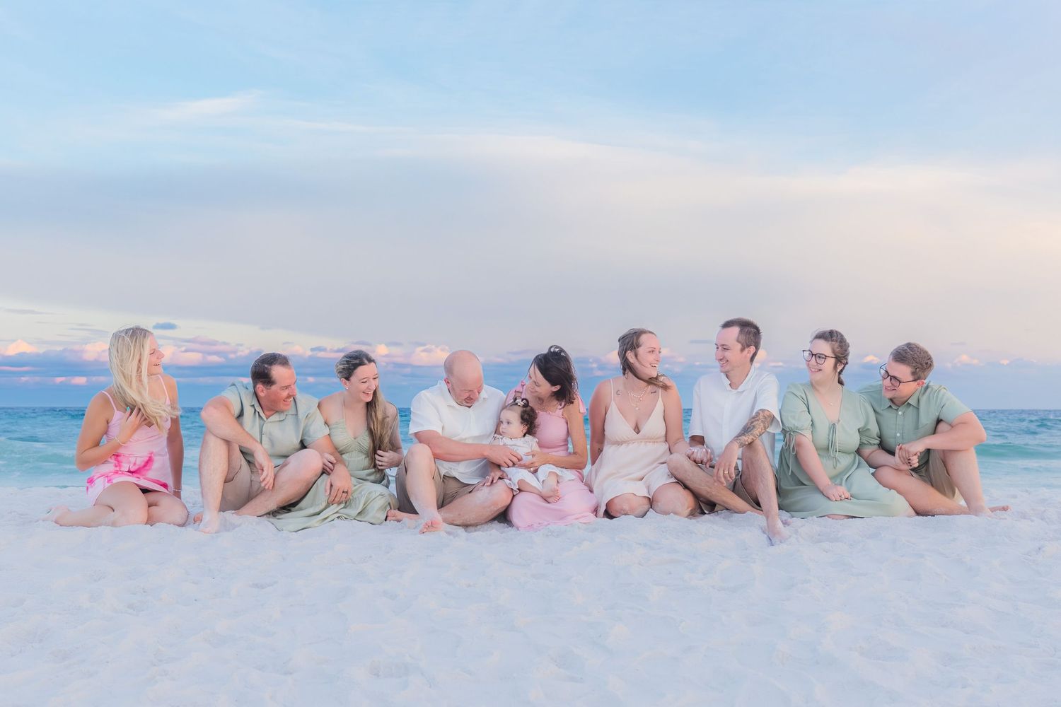 Large family group sitting together on a white sand beach at sunset, wearing coordinated green and pink outfits.