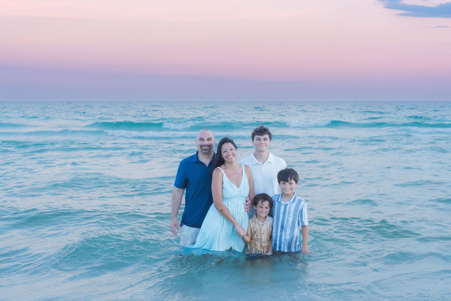 Family of five posing together in shallow ocean water at sunset with a pink and blue sky backdrop.
