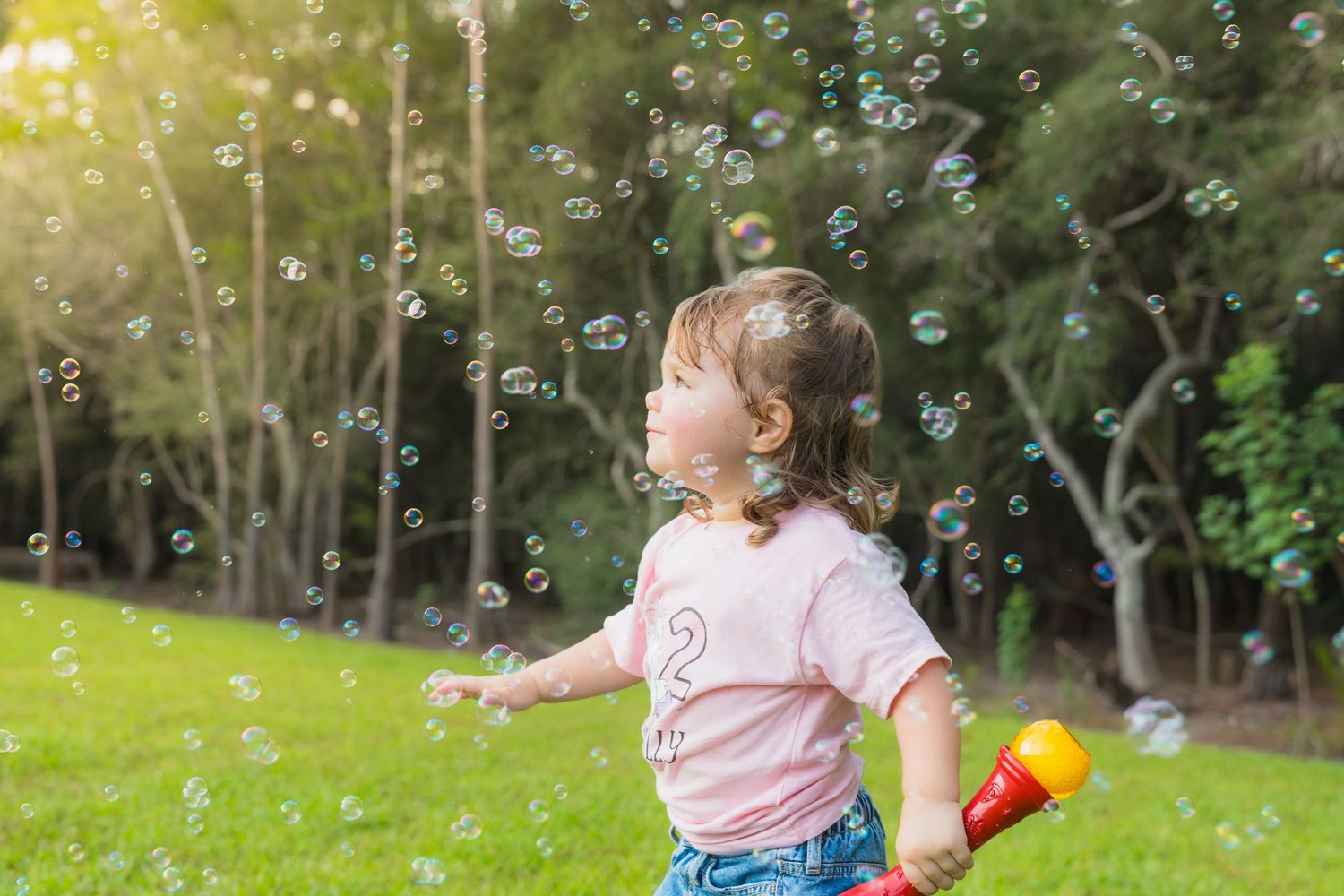A young girl in a pink shirt plays with a bubble gun outdoors, surrounded by floating soap bubbles in a green park.