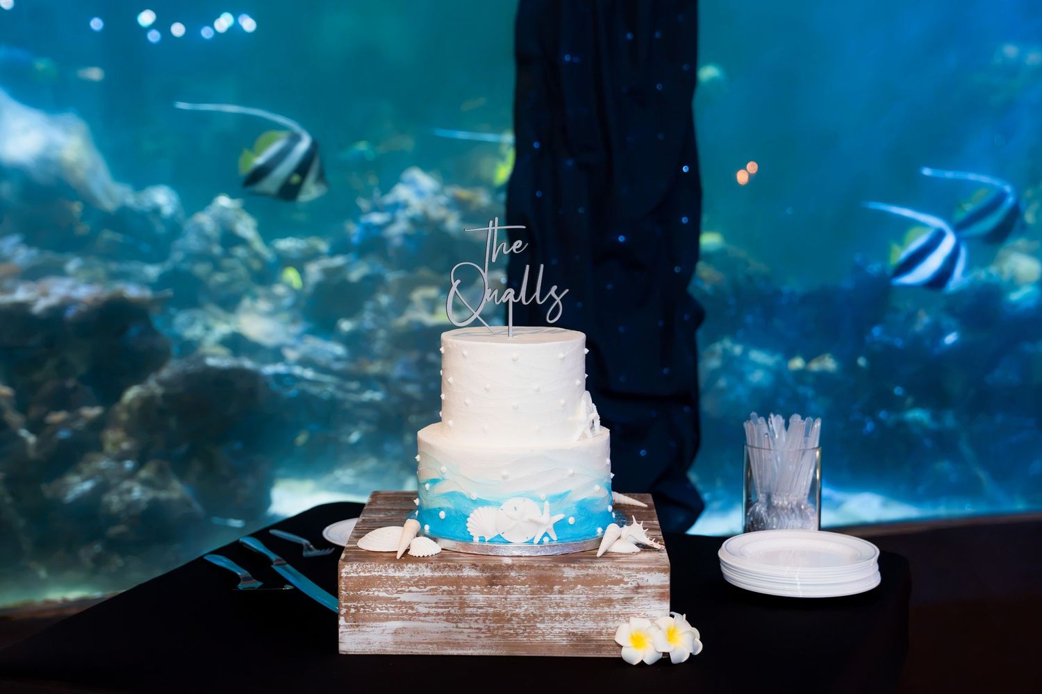 Three-tier wedding cake with blue ocean theme displayed in front of large aquarium fish tank.