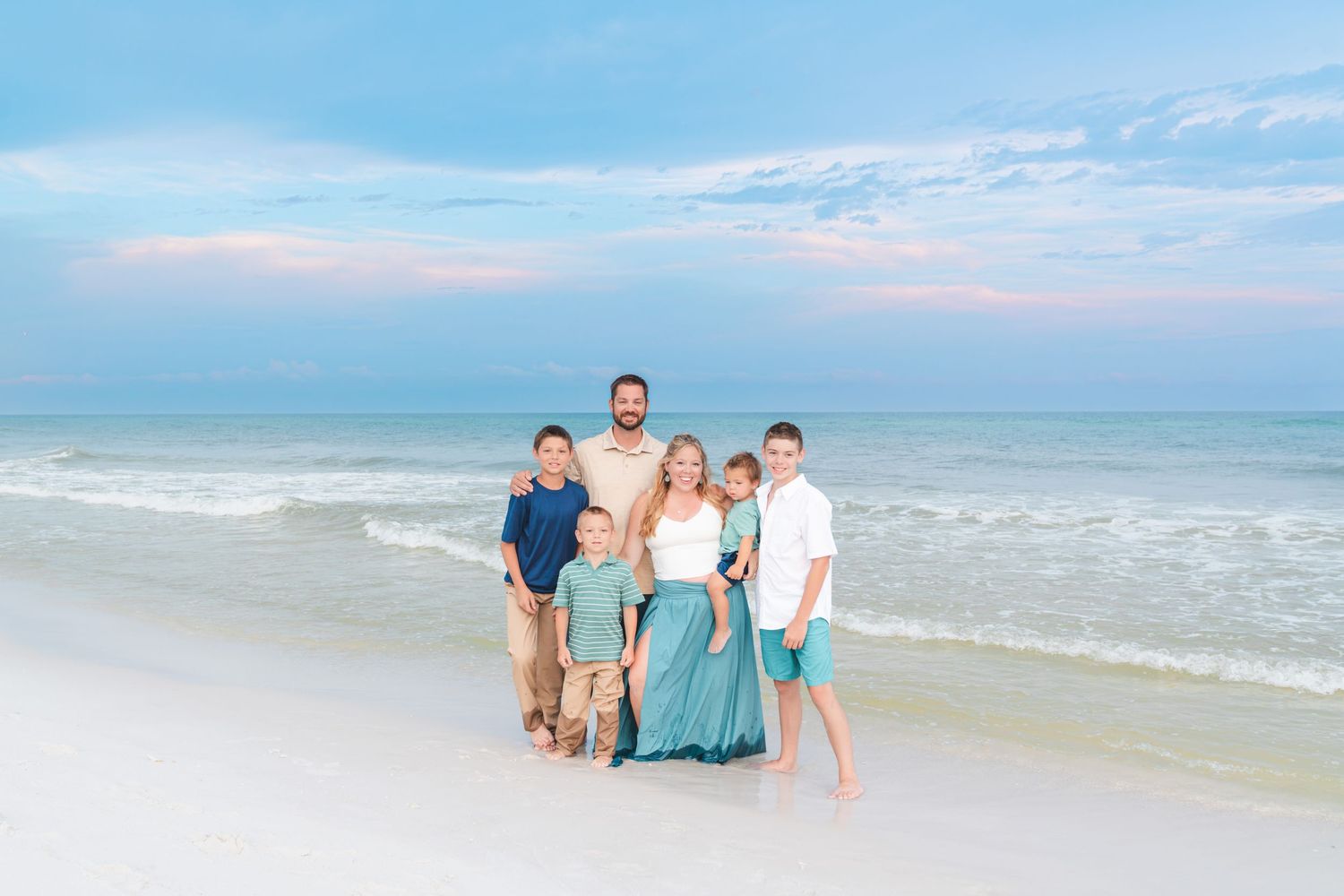 Family of six posing together on a white sand beach at sunset with turquoise ocean waves behind them.