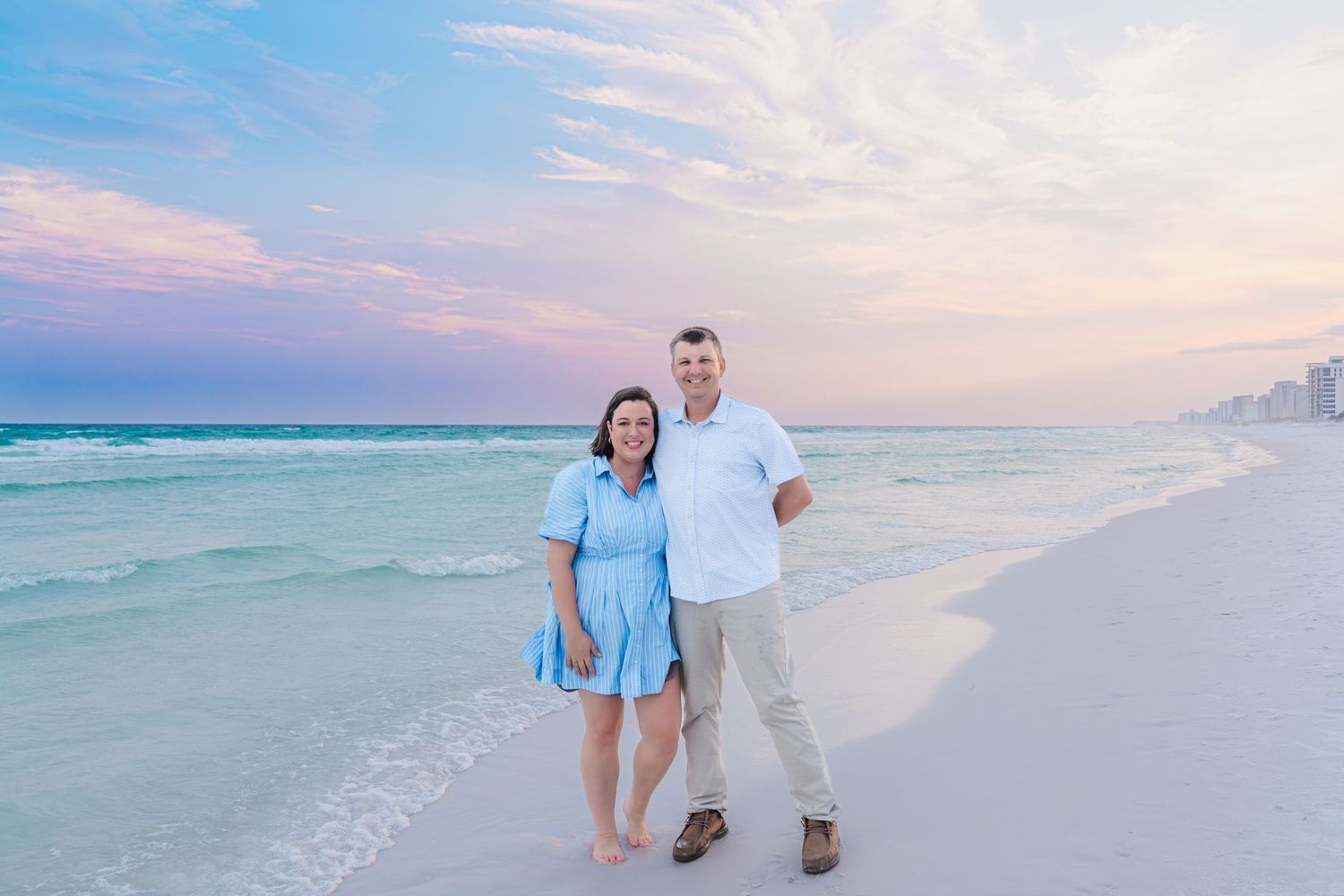 Couple smiling together on a white sand beach at sunset with turquoise waves and a colorful pink and blue sky.